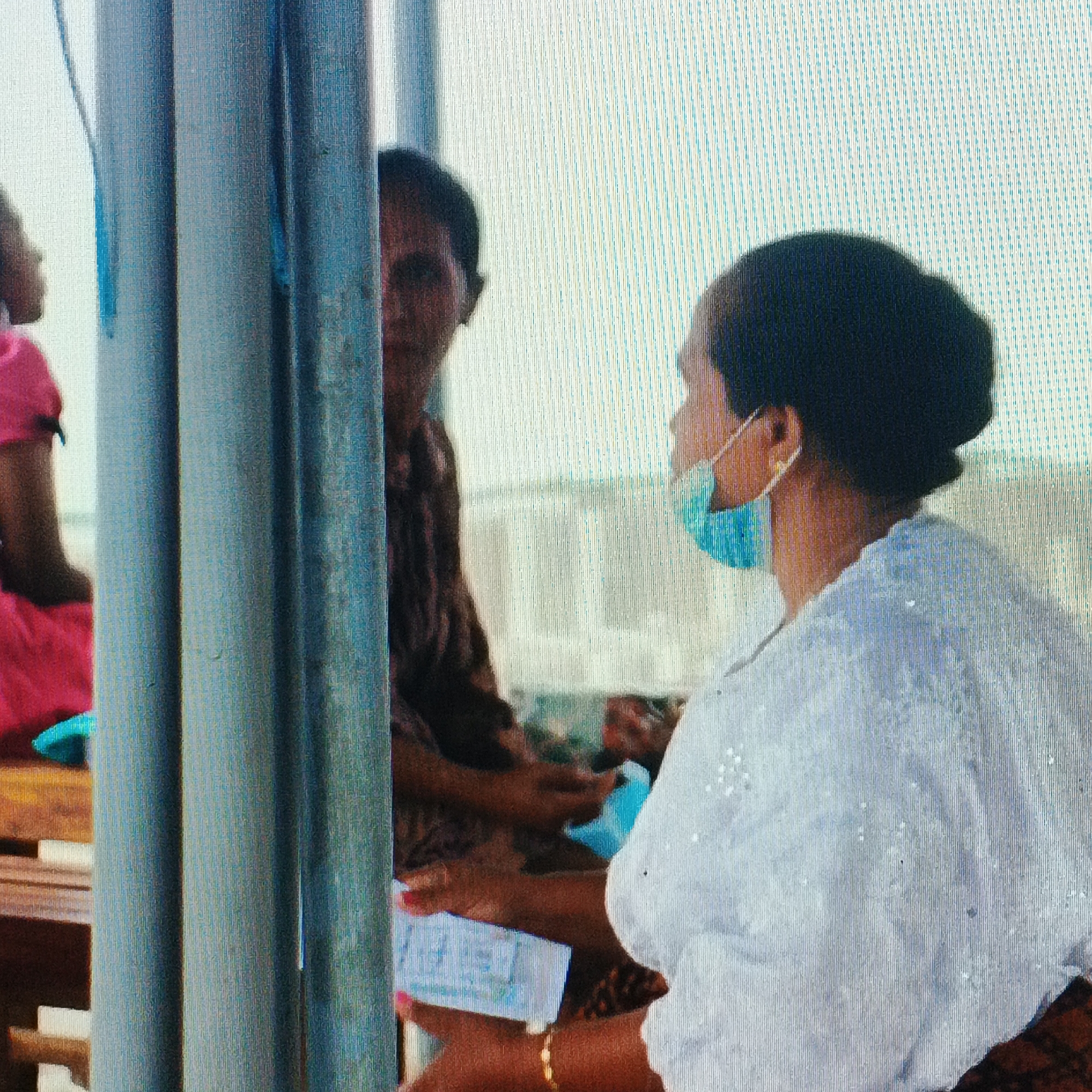 The image shows a close-up of two women interacting, with a portion of a pink-clad arm and shoulder visible on the far left. The woman on the right, facing left, is wearing a light blue face mask and a white garment adorned with intricate patterns and possibly embellishments. Her hair is styled in a dark bun. She is holding something white and rectangular in her hands. To her left, another woman, partially obscured by a blue-grey vertical pole, is seen from the chest up, wearing a dark, patterned garment. Her right hand is visible, holding a white rectangular object, possibly a ticket or paper, with some pink on her fingernails. A thin gold bracelet is visible on her wrist. The background is a soft, out-of-focus expanse of white and light blue vertical lines, suggesting an ambient light source or a patterned wall/window. The image appears to be taken indoors or in a sheltered outdoor area, given the presence of the vertical poles and the soft lighting. The context of Ende, Indonesia, is not directly evident from the visual information presented in this cropped image, as there are no specific landmarks or cultural indicators that definitively place it in that location.