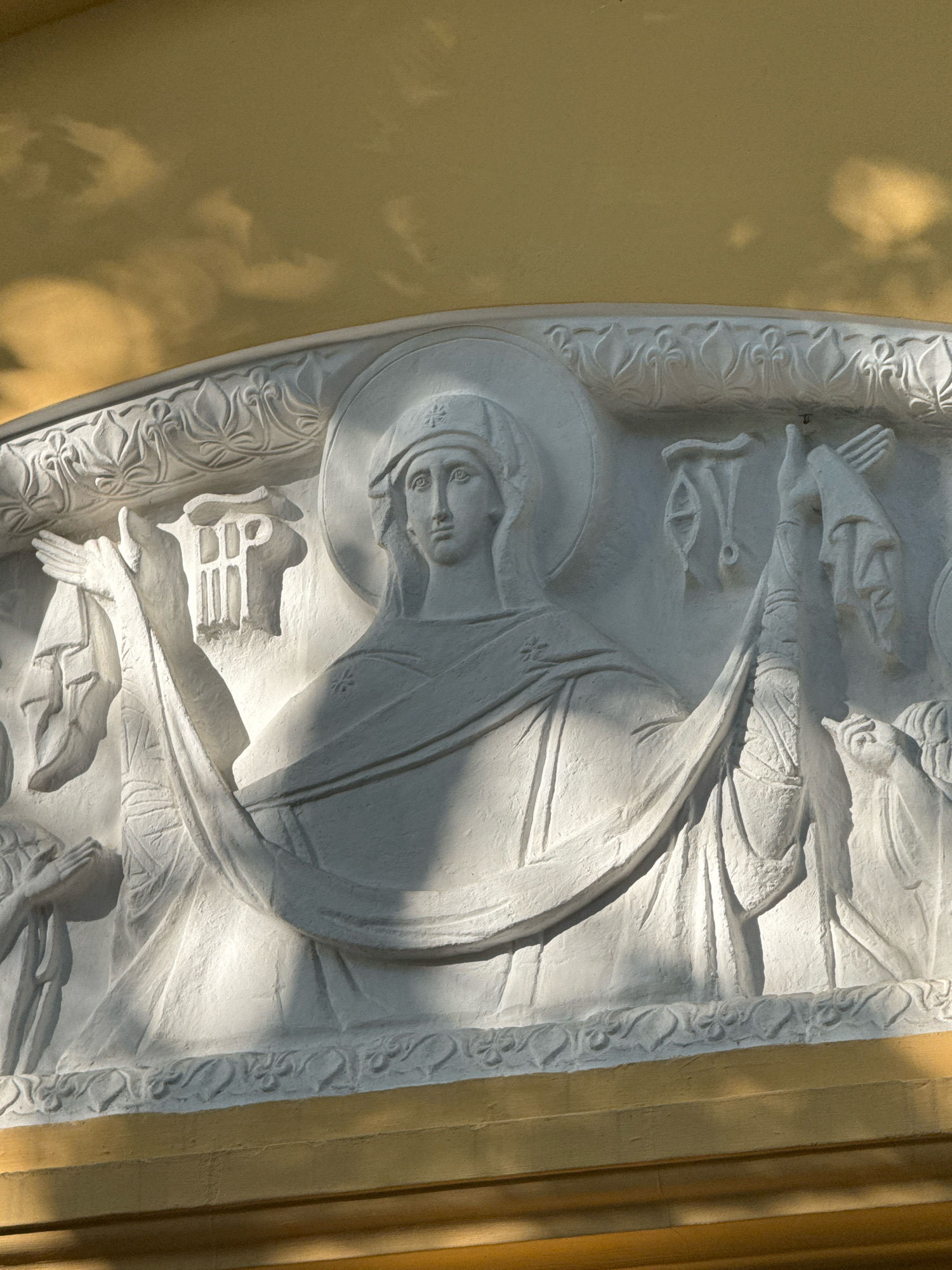 The image shows a relief sculpture depicting a central female figure with a halo, dressed in classical robes. Her arms are extended, and she holds a draped cloth. To her left, another figure is partially visible, with hands clasped in prayer. To her right, there are other symbolic elements and figures. The sculpture is made of white stone and is mounted on a building with a yellow, textured surface below. The background above is a soft yellow-ochre, possibly a wall or ceiling, with dappled light suggesting shadows. The sculpture is located on the facade of the Saint Petersburg Synodal Building (also known as the Building of the Holy Synod and Main Spiritual Directorate), situated at 6 Square of the Revolution. The architectural style is Neoclassical, and the relief likely represents a religious or allegorical theme.
