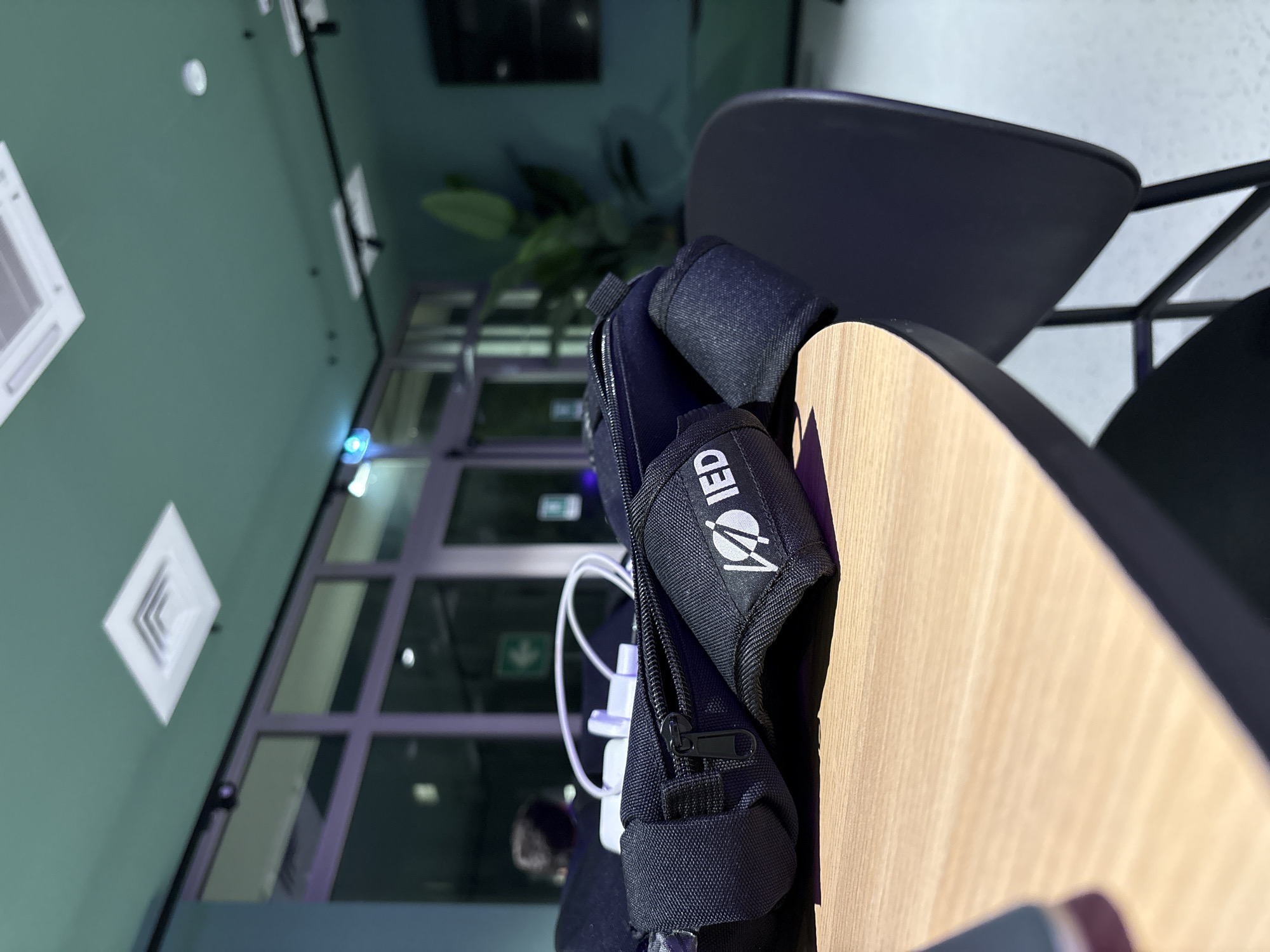 A close-up, low-angle shot focuses on a black fabric pouch with a zipper and a white logo that reads "IED" on a light wood table. White cables are visible within the pouch. In the background, large windows with purple lighting and reflections are seen. A dark chair is partially visible to the right, and further in the background, a large green plant and a television screen are present. The ceiling, painted teal, features air vents and light fixtures. The scene appears to be an indoor public space, possibly a cafe or co-working area, located in Novate Milanese, Italy. There are no discernible people in focus, and no specific actions or interactions are taking place.