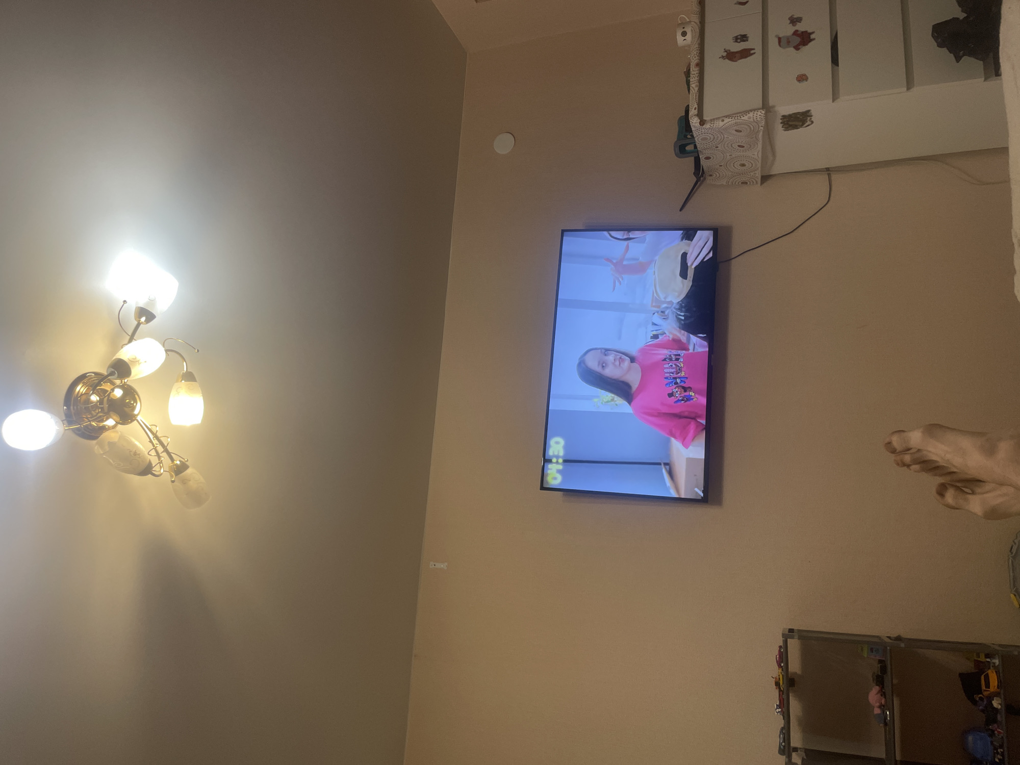 The image shows an indoor scene, likely a living room, with a television mounted on the wall. The TV displays a program with a young girl wearing a pink t-shirt. The room is dimly lit, with a ceiling fan with multiple lights visible at the top of the image. To the left of the television, a grey shelving unit holds various items, including toy cars. To the right of the television, a white dresser with drawers is decorated with Christmas-themed stickers. In the foreground, a pair of feet are visible, suggesting someone is sitting or lying down, watching the television. The walls are painted a neutral color. The location is identified as Laishevo, Russia.