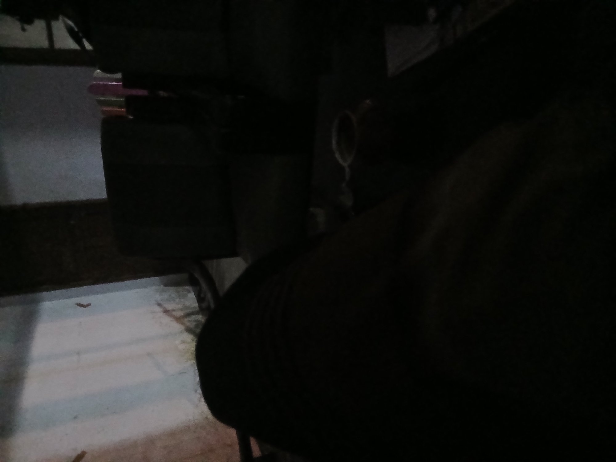 The image appears to be taken in a dimly lit room, possibly a living room or lounge area. In the foreground, a dark mass, likely a person's leg covered in dark clothing, dominates the lower half of the frame. The texture suggests fabric. To the right of the leg, on a dark surface, there is a light-colored ring, possibly from a cup or mug, with a dark liquid inside. Further in the background, there are dark, indistinct shapes that could be furniture, such as sofas or chairs. A wall is visible on the left, illuminated by a faint light source, showing vertical lines that might be part of the wall's structure or a curtain. The overall impression is one of a casual, indoor setting, possibly at night or in low-light conditions. The specific location within Tenggarang, Indonesia, cannot be determined from the visual information provided.