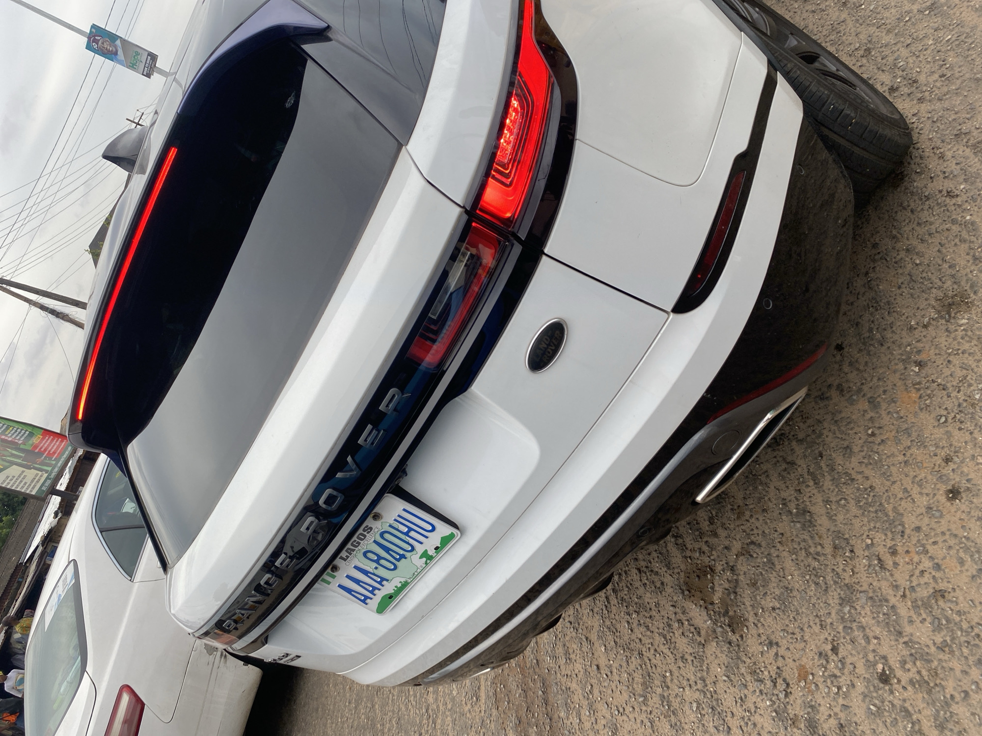 The image captures the rear view of a white Range Rover, likely a Range Rover Evoque, parked on what appears to be a street or unpaved lot in Agege, Lagos, Nigeria. The vehicle features sleek black accents, including a black roof and rear bumper, contrasting with its white body. Its distinctive LED taillights are illuminated bright red, and the "RANGE ROVER" lettering is visible across the tailgate, along with a "LAND ROVER" emblem on the right side.
A Nigerian license plate, clearly displaying "LAGOS," "CENTRE OF EXCELLENCE," and the alphanumeric code "AAA-840HU," is mounted at the center of the rear. To the right of the car, a black tire leans against its bumper. Another white car, a sedan, is partially visible behind the Range Rover, suggesting a crowded parking or street environment.
The ground beneath the vehicles is a rough mixture of dirt and gravel. In the background, against an overcast sky, overhead wires crisscross, and a banner or billboard is visible with partial text including "Hope" and "DOOR TO DOOR." The overall lighting suggests a cloudy day, with no harsh shadows.