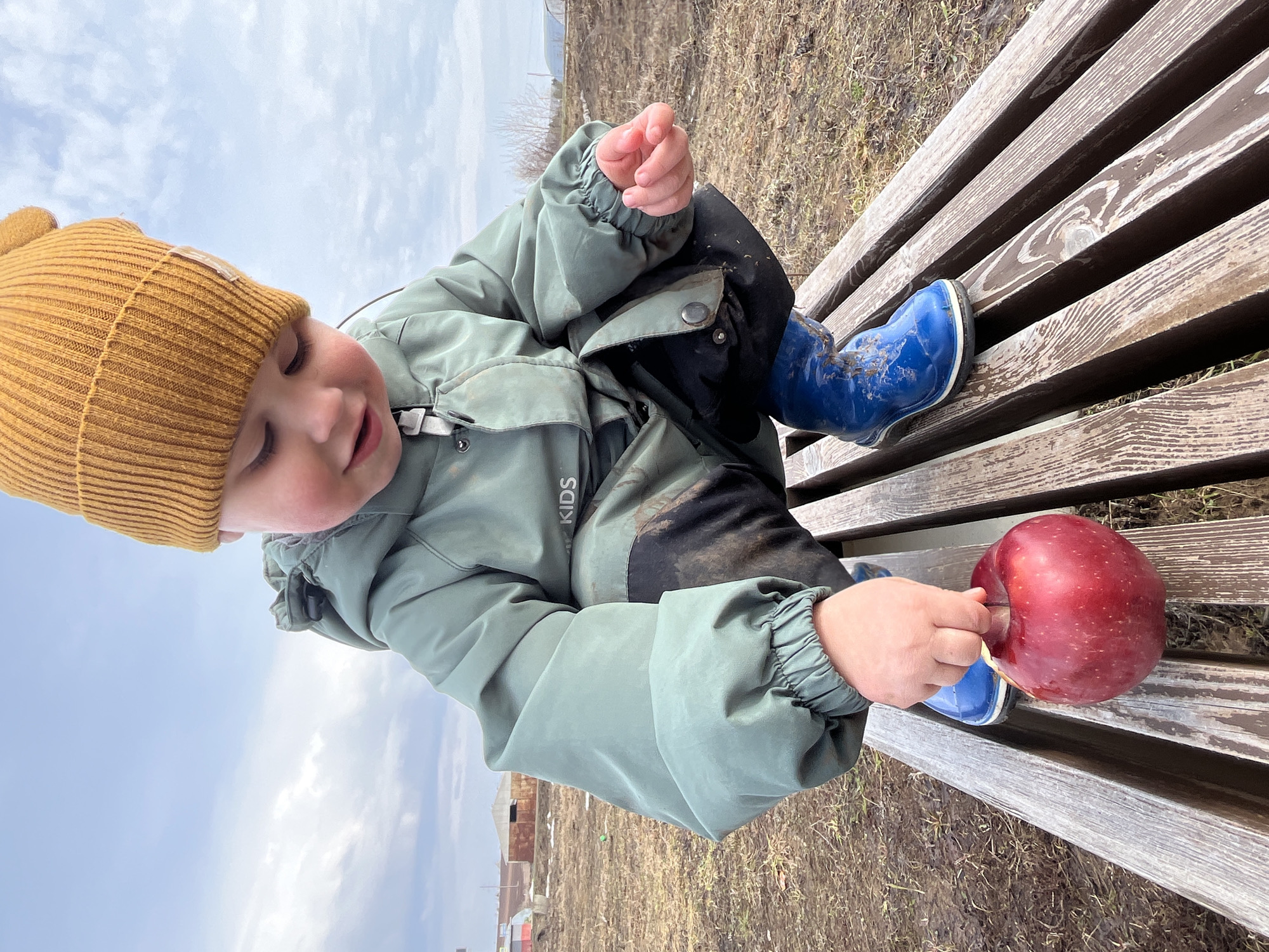 A child is sitting on a wooden bench outdoors. The child is wearing a mustard-colored beanie, a light green jacket, black pants, and blue rain boots. The child is holding a red apple. The background shows a field with sparse vegetation and a cloudy sky. The location is Павловка, Russia.