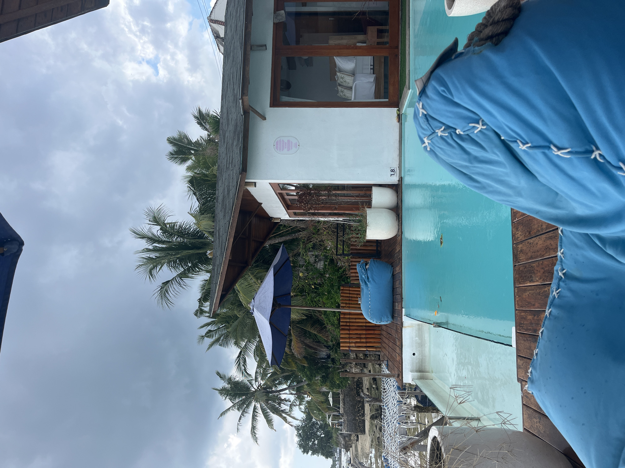 This image depicts a serene outdoor scene at a resort in Nusa Penida, Indonesia. In the foreground, a bright blue beanbag chair is partially visible, facing a swimming pool. The pool itself is a vibrant turquoise color, with a portion of it in the foreground appearing shallower and lighter, possibly due to sunlight or the pool's design. The pool is bordered by a wooden deck, upon which rests another blue beanbag chair and a large blue umbrella providing shade. Beyond the pool and deck, a white building with wooden-framed glass doors and windows is visible. The interior of the building reveals a bedroom setting with a neatly made bed adorned with pillows. Palm trees and lush green vegetation are scattered throughout the background, hinting at a tropical setting. In the distance, a glimpse of the ocean and rocky coastline can be seen, under a cloudy sky. The overall atmosphere is one of relaxation and tropical vacation.