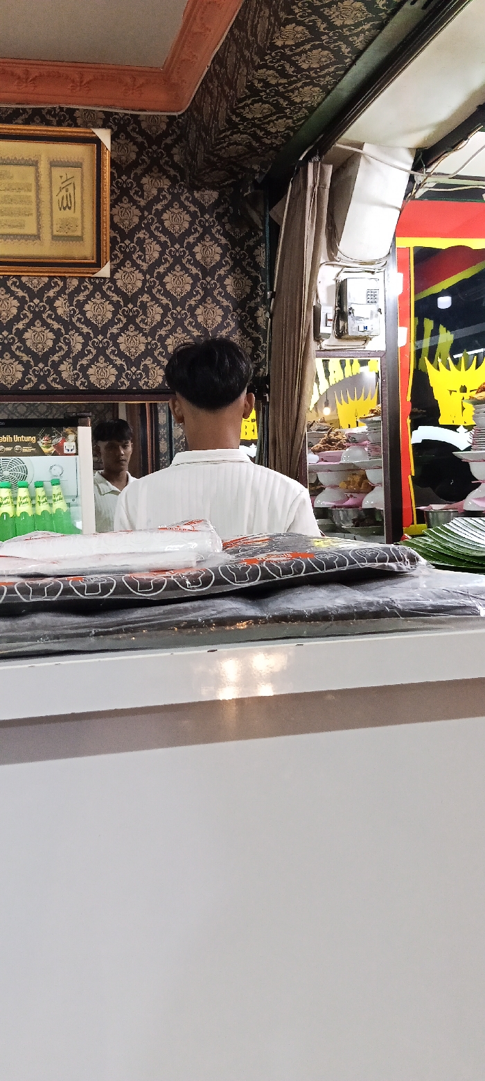 The scene takes place inside a shop in Kecamatan Kutawaringin, Indonesia, located at coordinates -6.988 latitude and 107.533 longitude. A man with short black hair, wearing a white shirt, is standing with his back to the camera facing a counter. He appears to be waiting to be served. In front of him is a white counter with a stack of black plastic bags on top. A man wearing a white shirt is partially visible in the background. A large framed picture hangs on the wall to the left of the counter. A refrigerator is visible on the left, with bottles of green liquid inside. In the background, a rack of goods can be seen on the right. The walls are decorated with floral wallpaper. There is an open doorway on the right side. It is unclear what the business is selling, but it is likely a food or drink establishment.