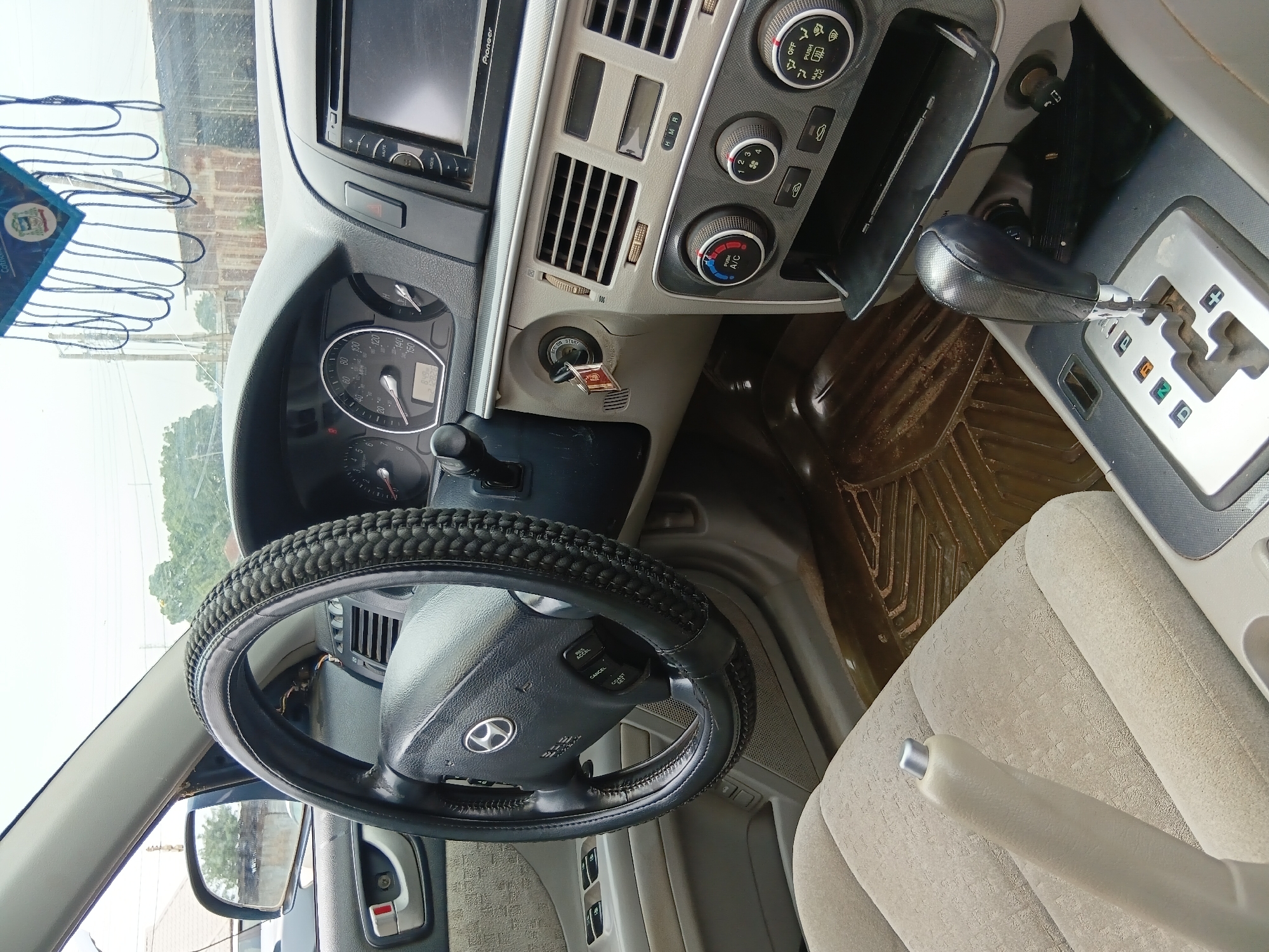 The image shows the interior of a Hyundai Sonata car, likely parked on a dusty road in Jos, Nigeria. The steering wheel has a black woven cover, and a car air freshener hangs from the rearview mirror. The dashboard features a standard instrument cluster, a Pioneer audio system, and climate control knobs. The gear shifter is in park position. The car's interior appears somewhat worn, with dusty floor mats and seat fabrics. The exterior view through the windows suggests an overcast sky and typical urban or semi-urban scenery of Jos, with buildings and vegetation visible.