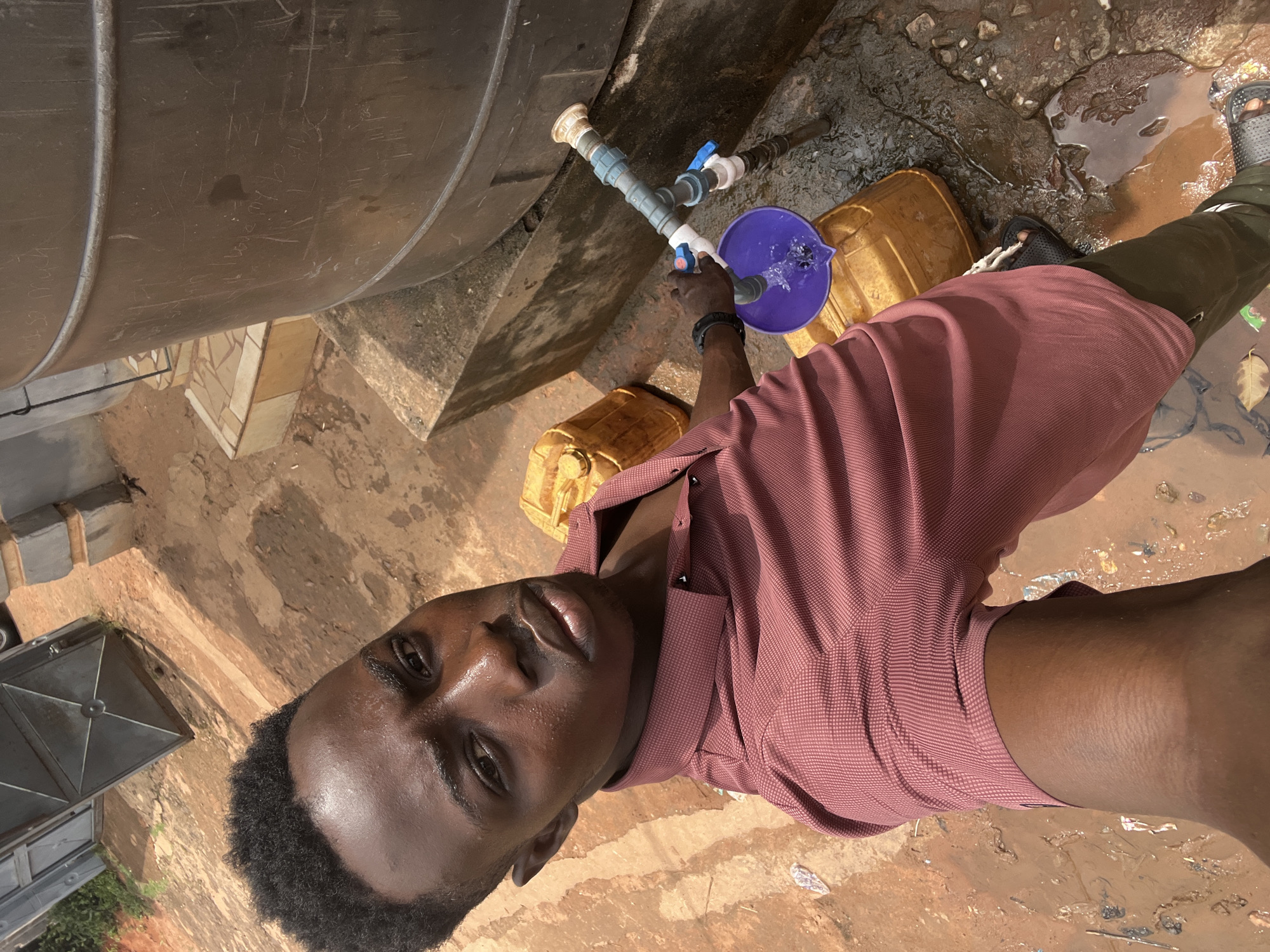 A young, dark-skinned man is shown from the waist up, holding a blue plastic bowl under a PVC pipe coming out of a large, black water tank. Water is pouring from the pipe into the bowl. He is wearing a maroon, short-sleeved, button-down shirt and dark trousers. He is looking at the camera, which is held at an angle. The ground is muddy and littered. There are yellow jerrycans near the water tank. The scene appears to be located outdoors. The location is Obosi, Nigeria.