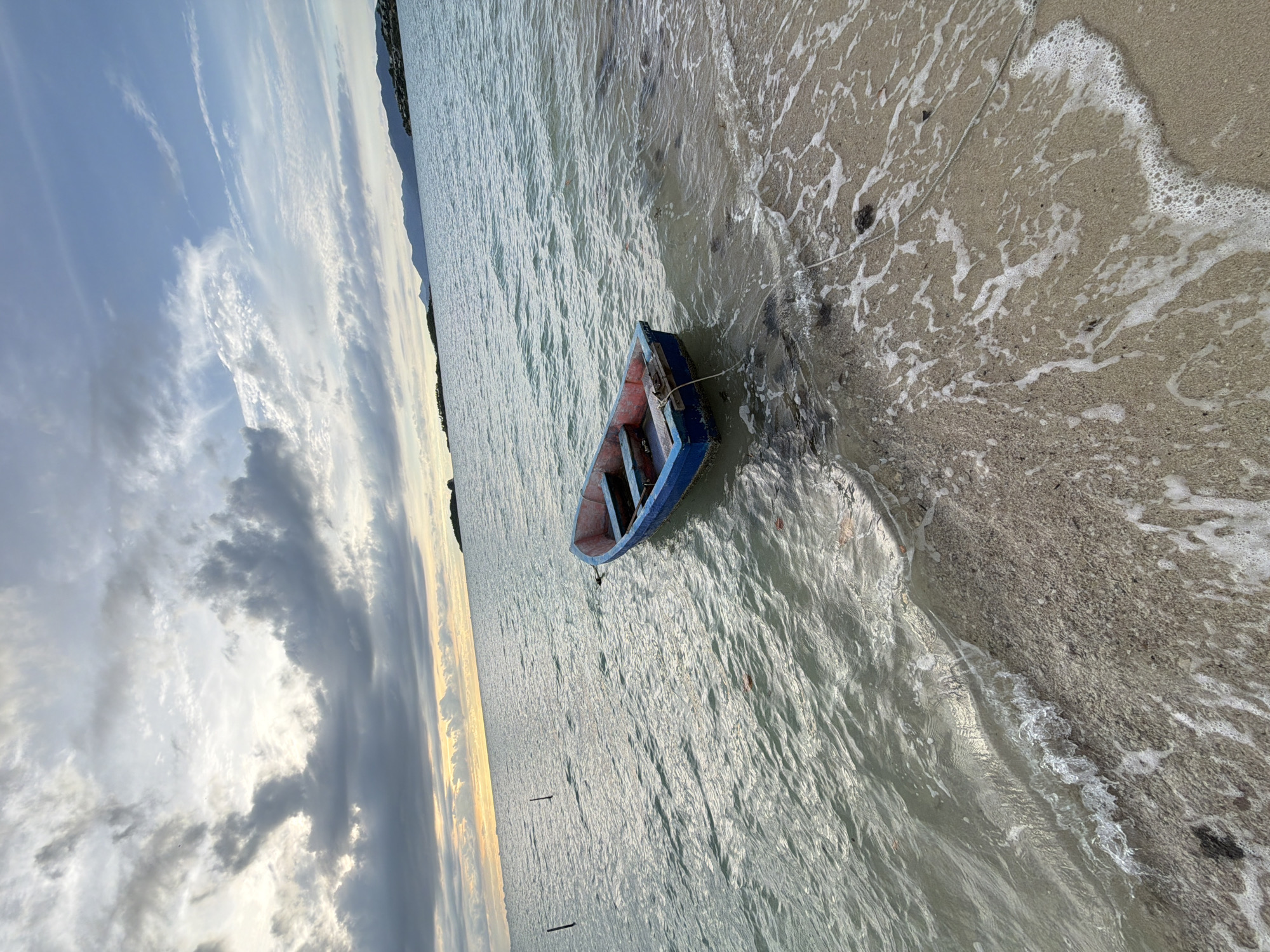 In Baan Plai Laem, Thailand, a small, blue and red rowboat rests partially beached on a sandy shore. The boat, unoccupied, is tethered by a rope to an unseen point, with its stern facing the water. Gentle waves lap at the sand, creating a foamy shoreline. The sea stretches out to the horizon, where a hazy line of distant islands and mountains can be seen. The sky above is a mix of dramatic clouds, with patches of sunlight breaking through, suggesting either dawn or dusk. The water is calm and reflects the muted light of the sky. There are no people visible in the image.