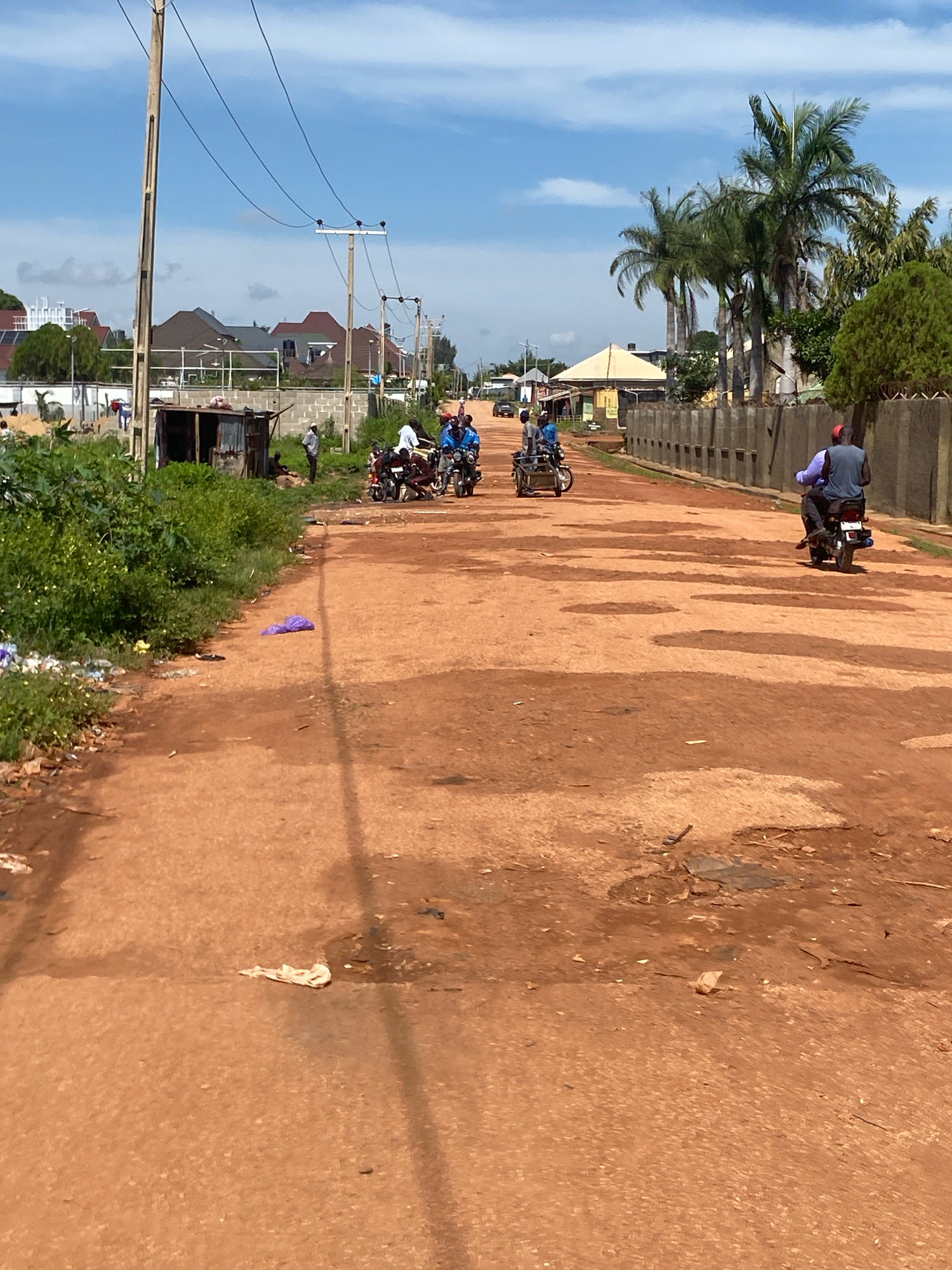 A dusty, unpaved road in Barden Yana, Nigeria, is lined with trees and buildings. Several people on motorcycles are traveling on the road, some with passengers. There is litter on the side of the road and in the foreground. The sky is blue with some clouds. The scene depicts a typical day in a Nigerian town, with transportation and daily life unfolding on the street.
