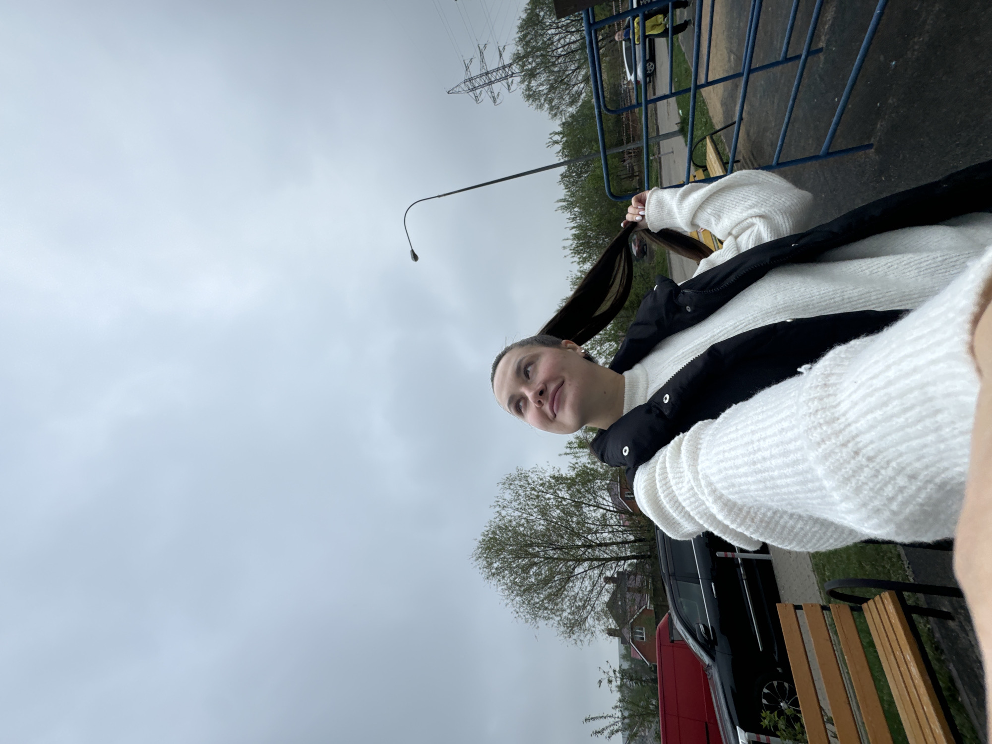 A young woman wearing a white sweater and black vest stands outdoors on an overcast day. She is holding her long, dark hair in her hands. In the background, there is a street with a street lamp and power lines overhead. There's also a blue metal fence, trees with sparse leaves, parked cars, and wooden benches. The location is identifiable as Akulovo in the Gp Kubinka region of Russia based on the architectural style of the buildings.