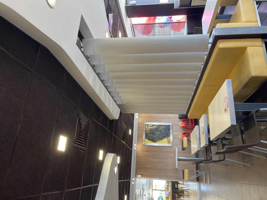 The image shows a McDonald's restaurant interior in Hadjidimitrovo, Bulgaria. The image is taken from the seating area, looking towards a booth with a yellow cushioned bench and a set of white vertical blinds. The seating area has an assortment of tables and chairs. The floor is a beige tile. The far end of the restaurant is partially visible, featuring a wooden wall and large picture frame on the right wall. The ceiling is dark brown with recessed lighting. The image shows an empty restaurant with no customers or employees visible. There are two red stools visible to the left of the booth. The tables and chairs are clean and appear unused.