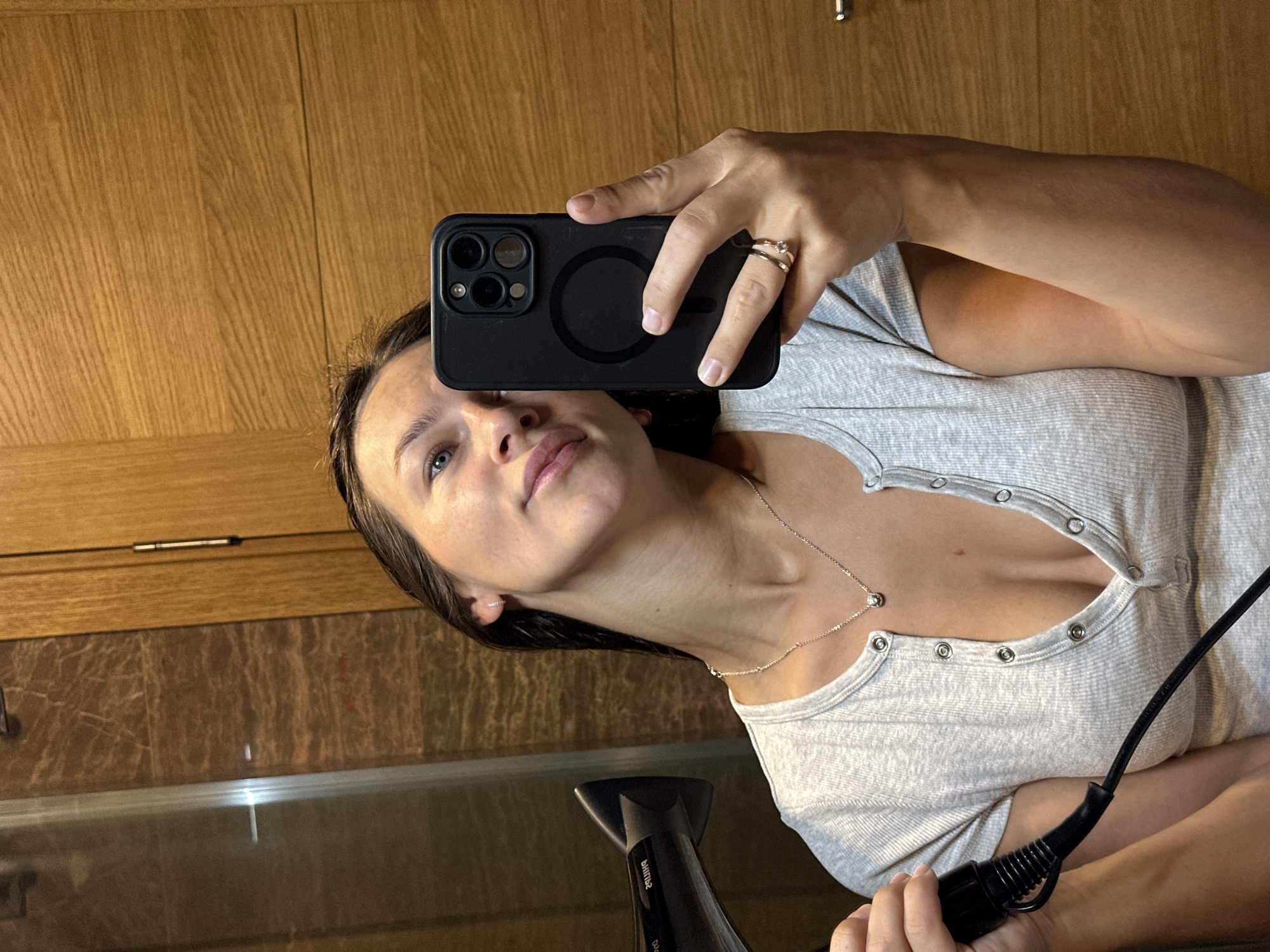 A woman is taking a mirror selfie while holding a hairdryer in her left hand. She is wearing a grey, short-sleeved, button-up shirt and a delicate necklace with a small pendant. A ring is visible on her right ring finger. The background appears to be a bathroom with marble-patterned tiles and a wooden door. The scene is indoors. The location is in the city of Donji Pobori, Montenegro.