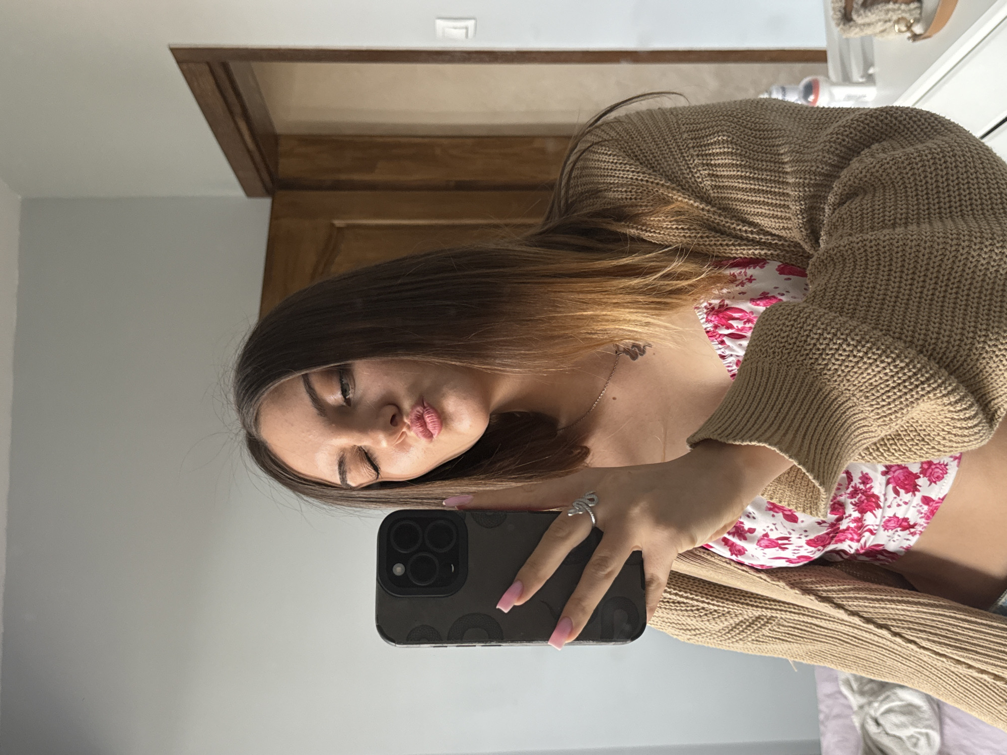 A selfie, taken indoors, depicts a young woman holding a dark-colored iPhone. She has long, brown hair with blonde highlights. She wears a floral print top under a tan-colored cardigan. Her fingernails are painted pink, and she wears a silver ring shaped like a snake on her ring finger. The background includes a gray wall and a portion of a wooden doorframe. The location is not discernible from the image and therefore cannot be identified as Sofia, Bulgaria.