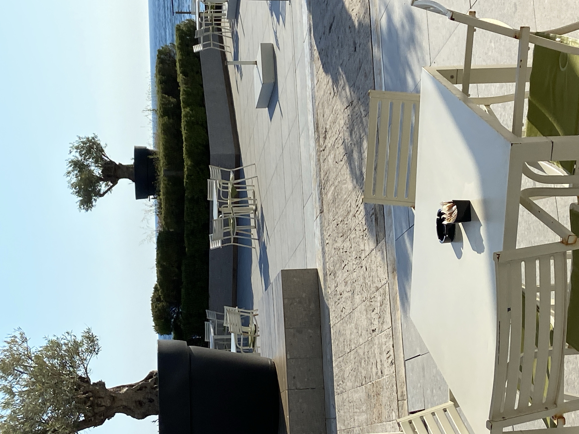 The image depicts an outdoor seating area in Petrovac na Moru, Montenegro. The scene includes several white tables and chairs, indicative of a cafe or restaurant setting. The foreground features a table with a container, possibly for straws or napkins, on it. In the background, there are green hedges and trees planted in large black planters, with glimpses of the sea visible beyond. The seating area is paved with light-colored tiles. The image is taken during the day, with bright sunlight casting shadows across the scene.