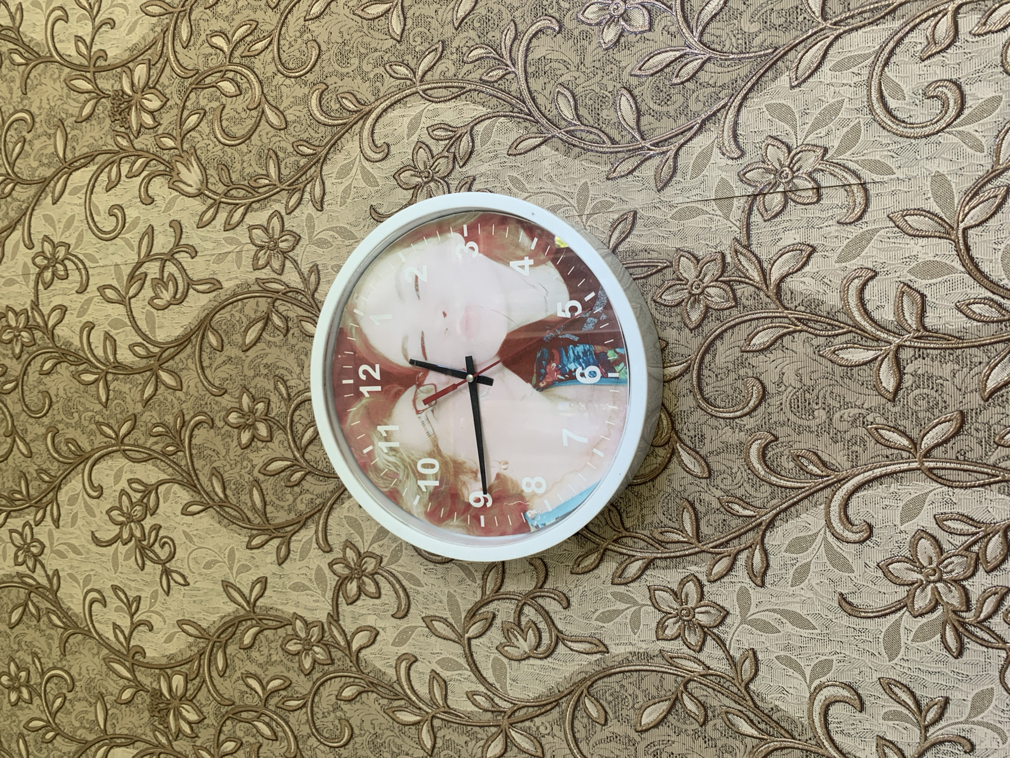 A white circular clock with a custom face featuring two young women is mounted on a wall with ornate floral wallpaper. The clock's hands are black, with the hour hand pointing slightly past the 10 and the minute hand pointing slightly past the 11. The wallpaper has a textured pattern of brown and beige floral and vine motifs. The scene appears to be indoors, likely a residential setting in Tashkent, Uzbekistan.