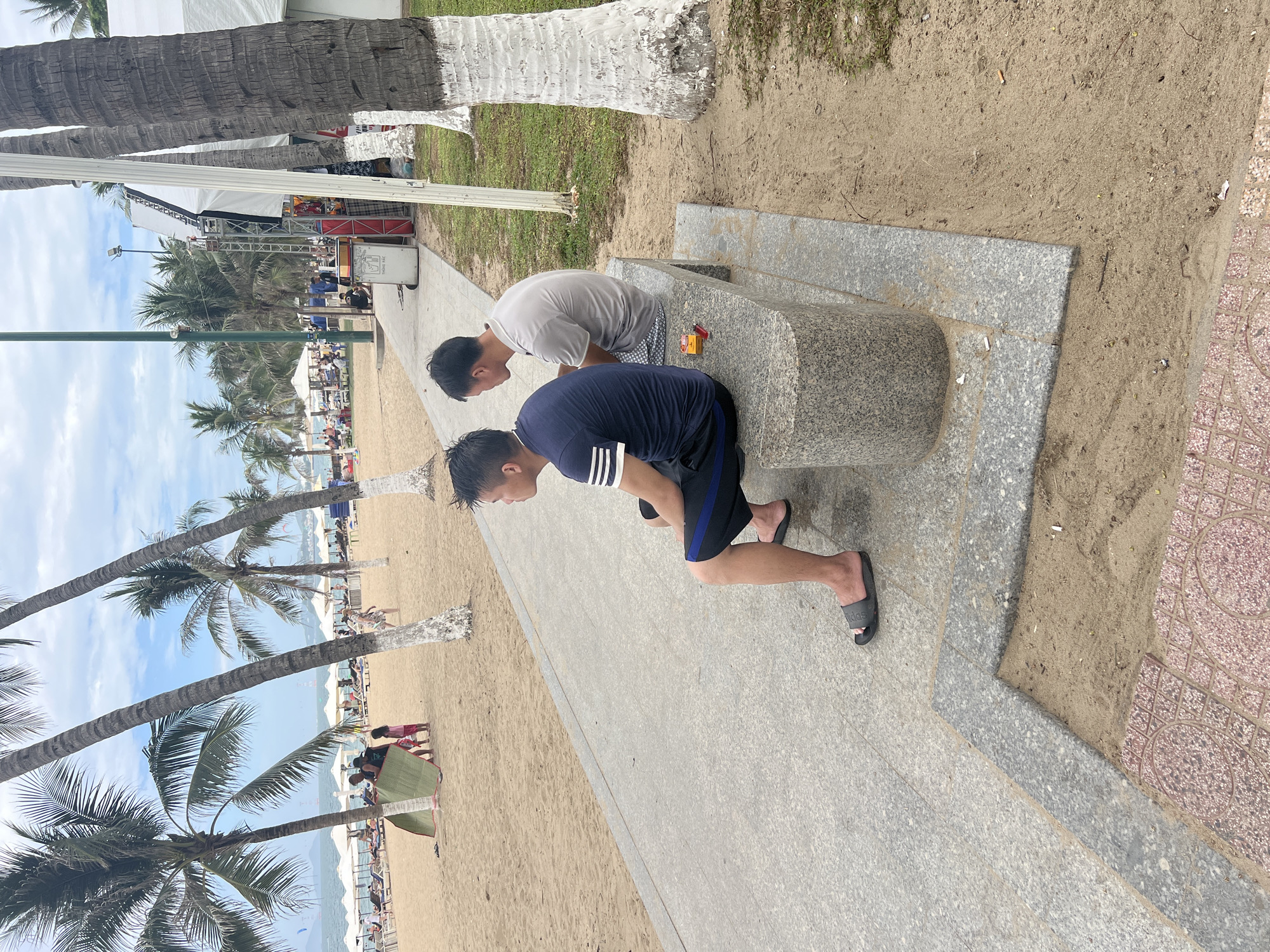 The image depicts a beachside promenade in Nha Trang, Vietnam. Two men are seated on a stone bench, facing away from the camera. The man on the left wears a dark blue t-shirt with white stripes on the sleeves and dark shorts. The man on the right wears a light gray t-shirt and checkered shorts. In front of them on the bench are a pack of cigarettes and a lighter. The beach is visible to the left of the promenade, with palm trees lining the sand. Beach umbrellas and lounge chairs are set up on the sand, and a few people are visible in the distance. A stage structure with lighting equipment is set up on the right side of the image, with tents and more palm trees behind it. The ground consists of sand and a paved walkway with granite borders. The overall scene suggests a public space designed for leisure activities by the sea.