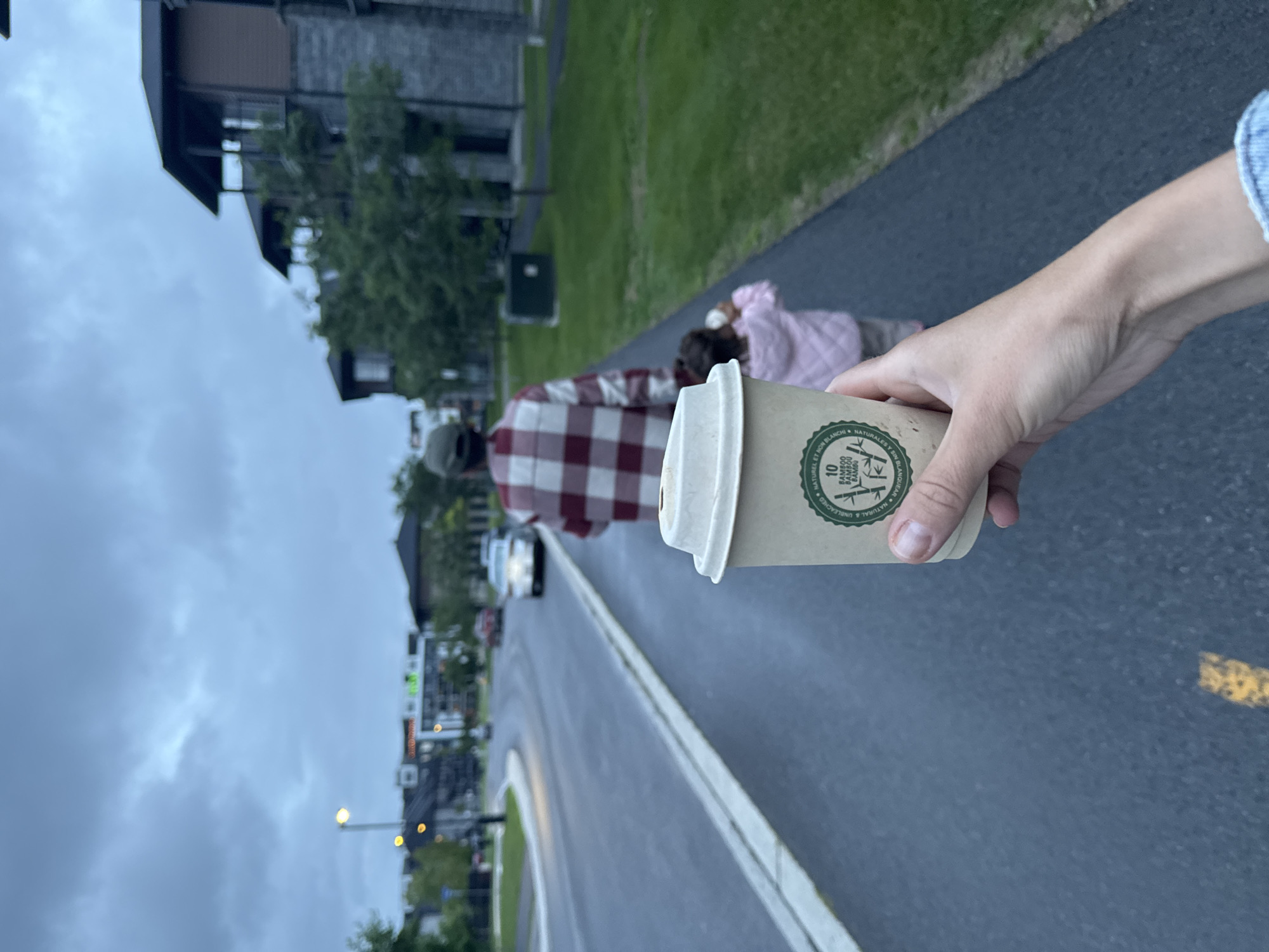 A first-person perspective shot shows a hand holding a disposable coffee cup as a man and child walk away on a paved pathway. The man wears a red and white plaid shirt and a green cap. The child, seen from the back, wears a pink jacket. The pathway runs alongside a road with a few cars and buildings in the background. The sky is overcast. The scene is likely in Gatineau, Canada, given the context. The coffee cup has a green circular logo with bamboo imagery and text that reads "10 BAMBOO BAMBOO BAMBOO BAMBOO NATUREL ET NON BLANCHI."