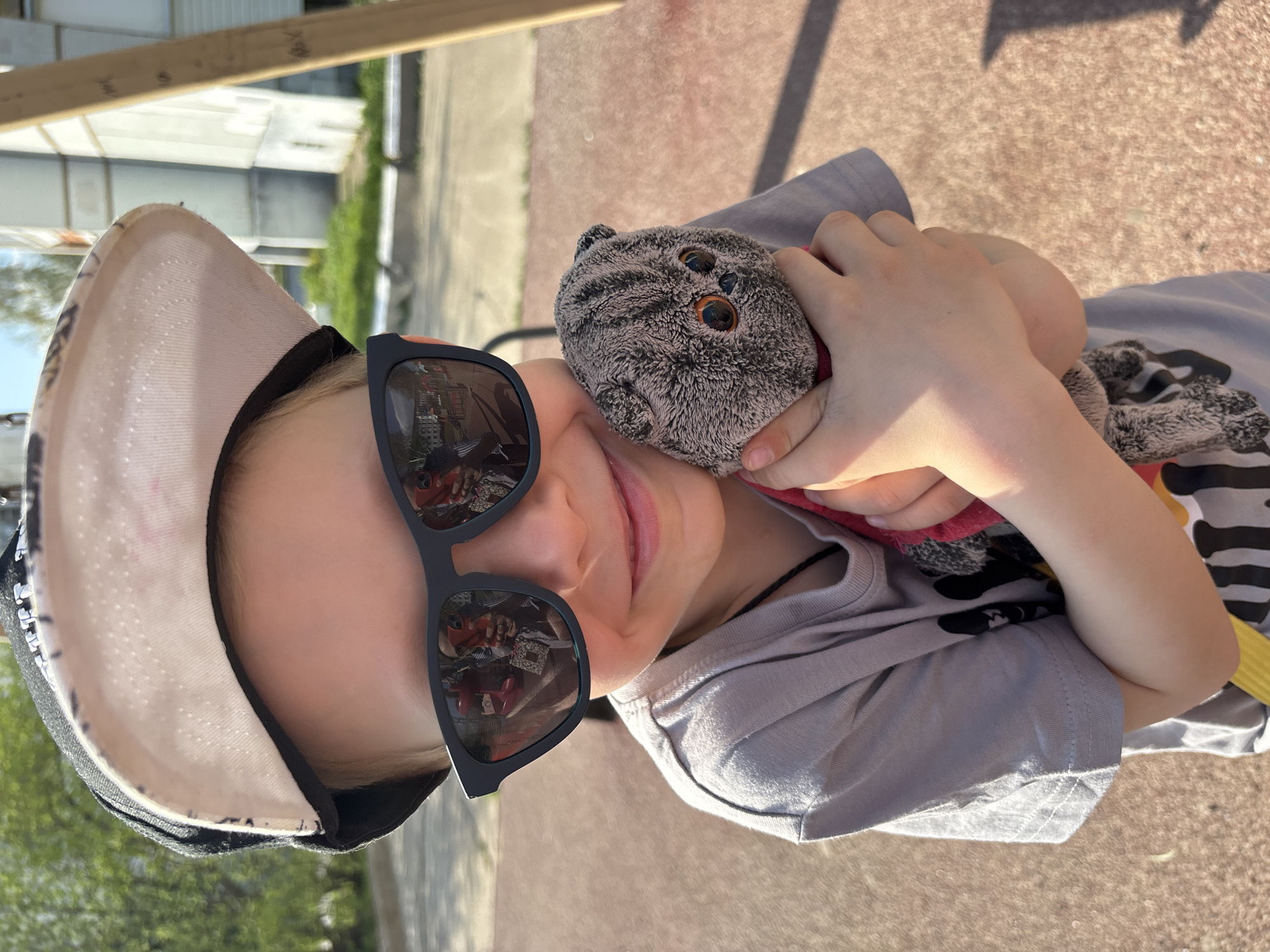 A child wearing a baseball cap and sunglasses holds a stuffed animal in an outdoor setting. The child is wearing a gray t-shirt. The background appears to be a playground or recreational area. The reflection in the sunglasses shows buildings and what might be a car, suggesting an urban environment. This is in the city of Chaikovsky, Russia.