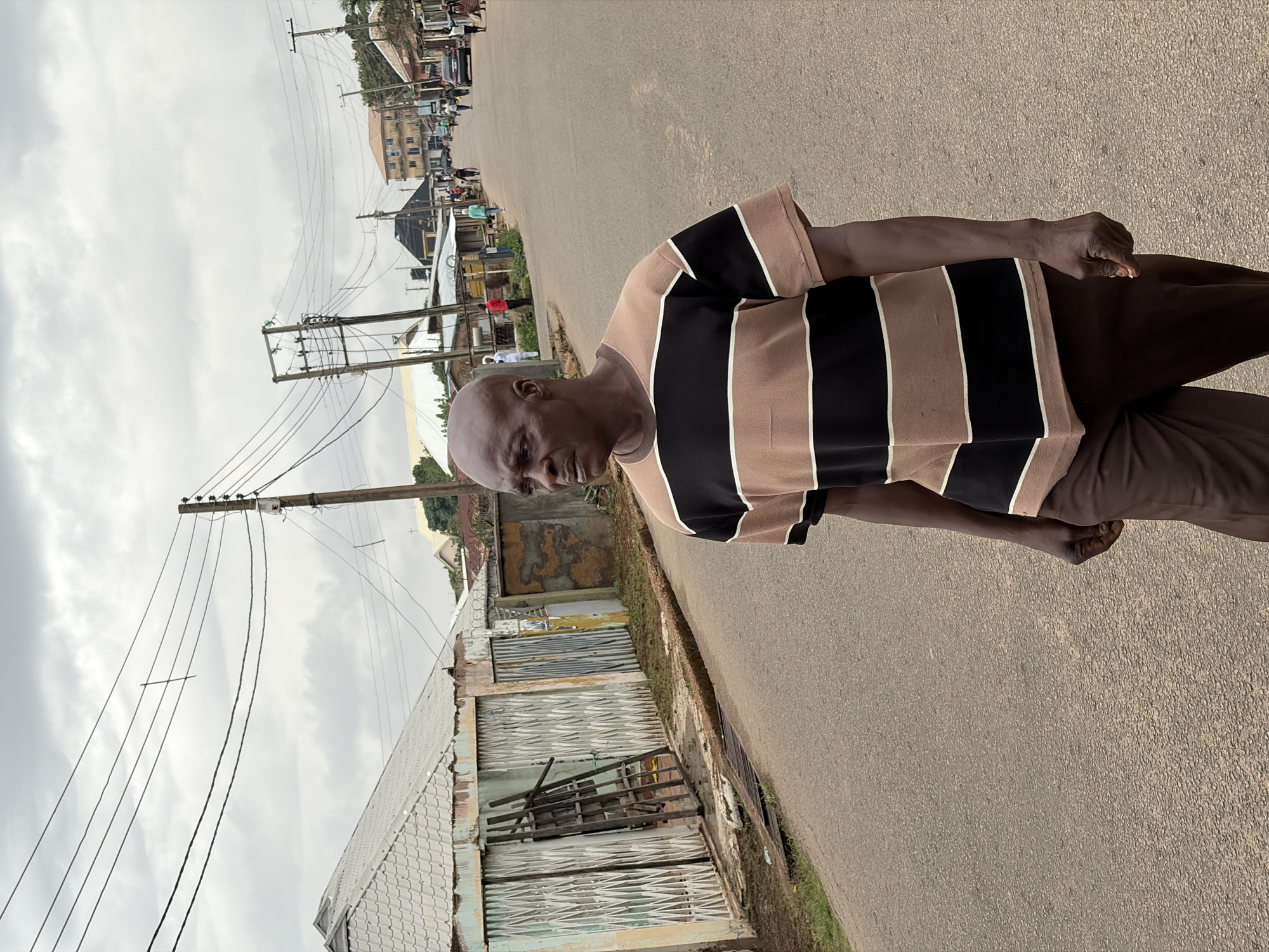 A bald, dark-skinned man stands on the asphalt of a road in Jos, Nigeria, looking towards the camera. He is wearing a tan and black striped shirt and brown pants. To his left, a building with a corrugated iron roof and dilapidated metal siding is visible. Power lines are strung overhead on wooden poles. In the distance, other buildings and vehicles are visible along the road, indicating an urban or semi-urban environment. The sky is overcast.