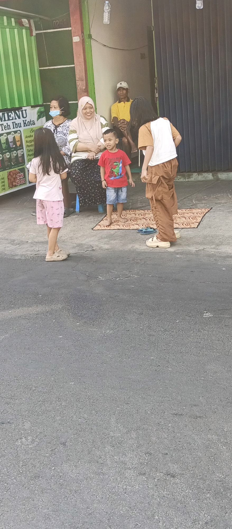 In front of a shop with a green striped awning and a dark metal roll-up door, a group of people are gathered on a paved surface. To the left, a menu board displays various drink options. Two adult women, one wearing a face mask and a patterned top, and another in a light-colored hijab and striped shirt, are seated. A young boy in a red t-shirt with a dinosaur graphic and blue shorts stands between them. To the right, a person with long dark hair wearing a white and tan shirt and brown cargo pants stands facing away from the camera. Another person with dark hair and a tan shirt is partially visible. A man in a yellow shirt and white cap sits in the background. A patterned mat is laid on the ground. The environment appears to be an outdoor setting in Malang, Indonesia, likely a street-side establishment.