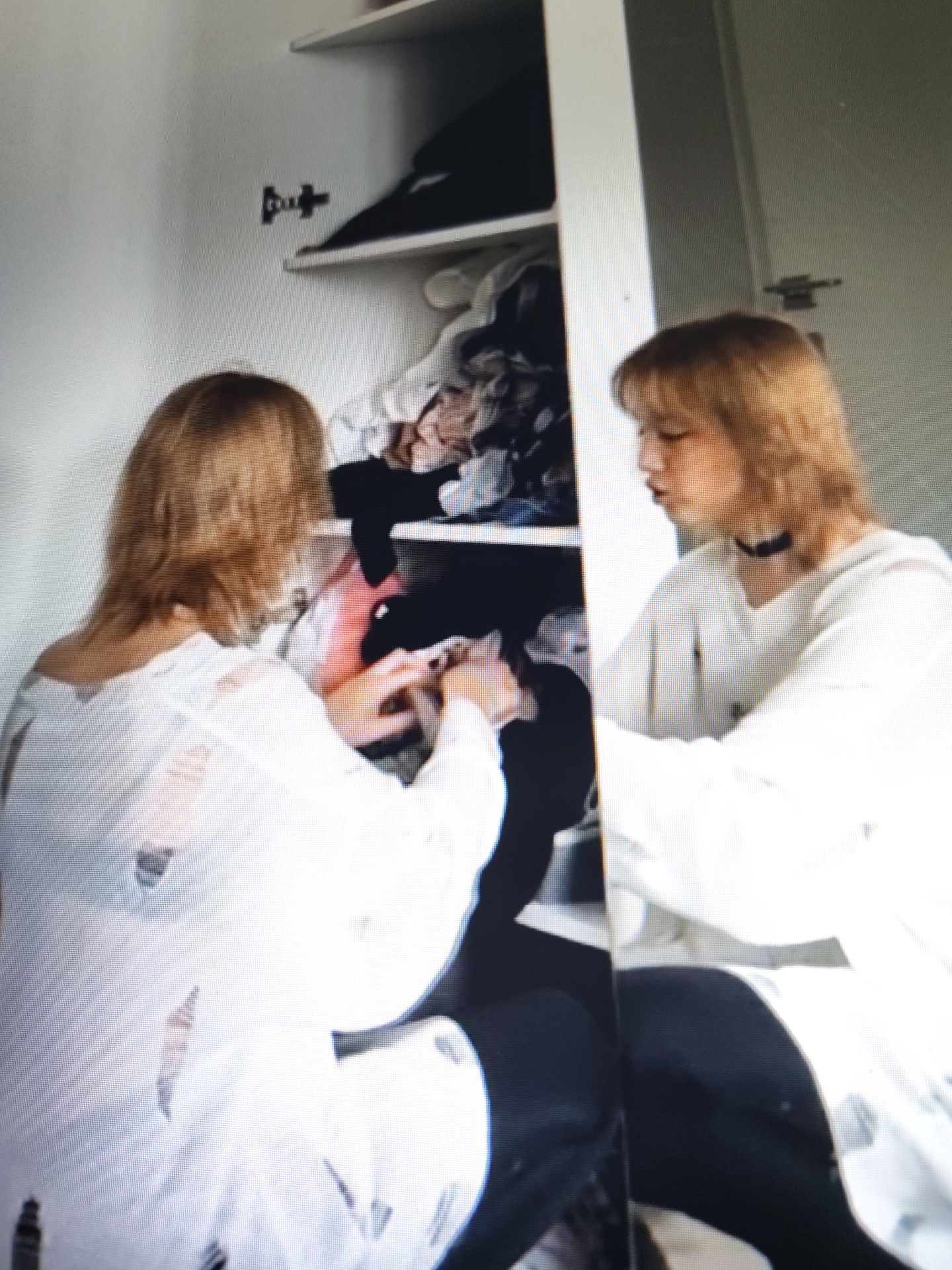 Two young individuals are seen in what appears to be a closet. One individual, with light brown hair, is facing away from the camera and is rummaging through clothes. They are wearing a ripped white shirt and black pants. The other individual, also with light brown hair, is in profile, facing left, and is wearing a white top and a black choker necklace. The closet shelves are stocked with various items of clothing. A mirror reflects the scene, showing the second individual again in profile. The image quality is poor, with noticeable pixelation. There are no visible indications of the specific location within Il’inskiy, Russia, in the provided image.