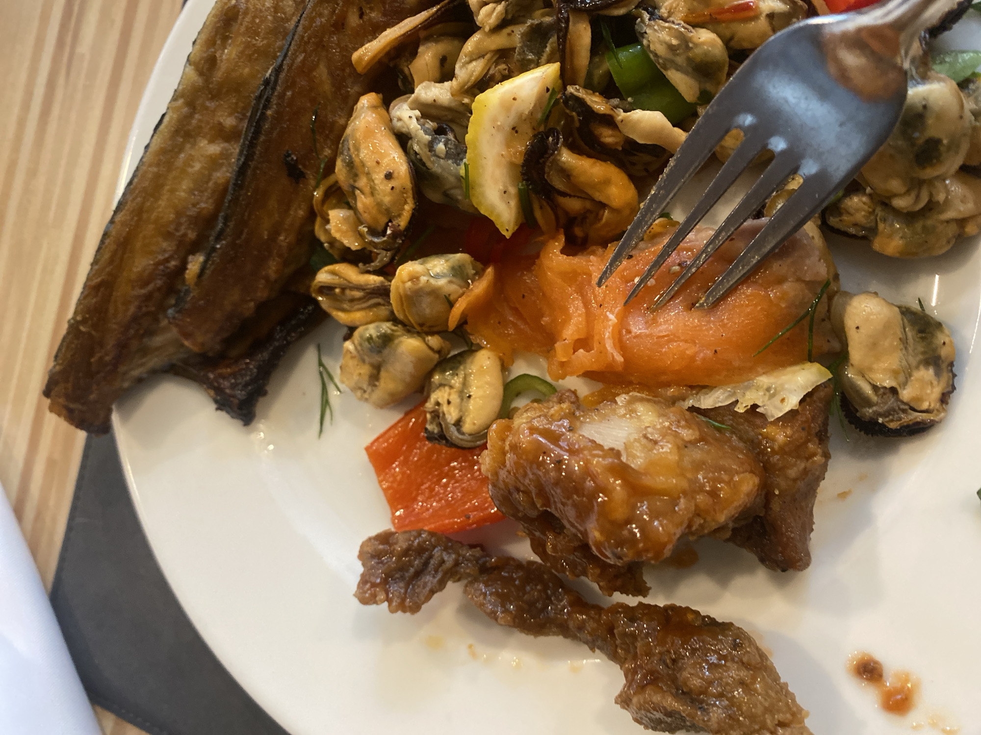 A plate of food is displayed on a wooden table with a white napkin to the left. The plate contains several dishes, including what appears to be fried eggplant strips, mussels, pieces of fried chicken with a sauce, smoked salmon, and red bell pepper. A fork is visible, resting on the salmon. The image is a close-up shot of the food, with the background blurred. There is no indication of the location being Aksu, Turkey.