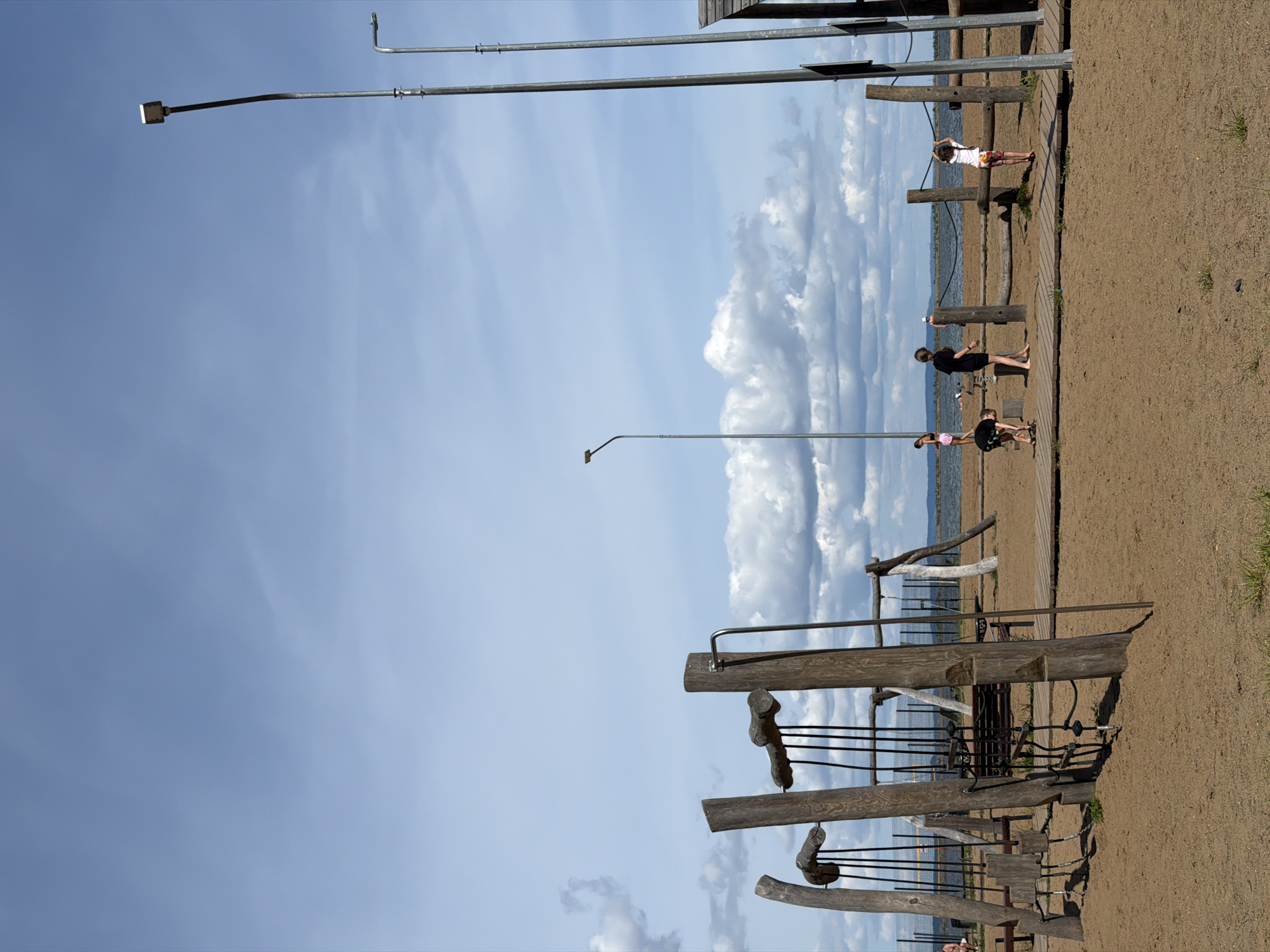 An outdoor playground with a beach area is depicted. In the foreground, a sandy surface is visible with a wooden playground structure. Several children are present. One child is bent over near the structure, another stands with their back to the camera, and a third is reaching up to a horizontal bar. In the background, a body of water is visible with a fence and more playground equipment. A few more people are scattered near the water. Tall metal lamp posts stand on either side of the scene, and clouds dot the bright blue sky. The scene appears to be located in the city of Chaikovsky, Russia, likely on the Kama River embankment.