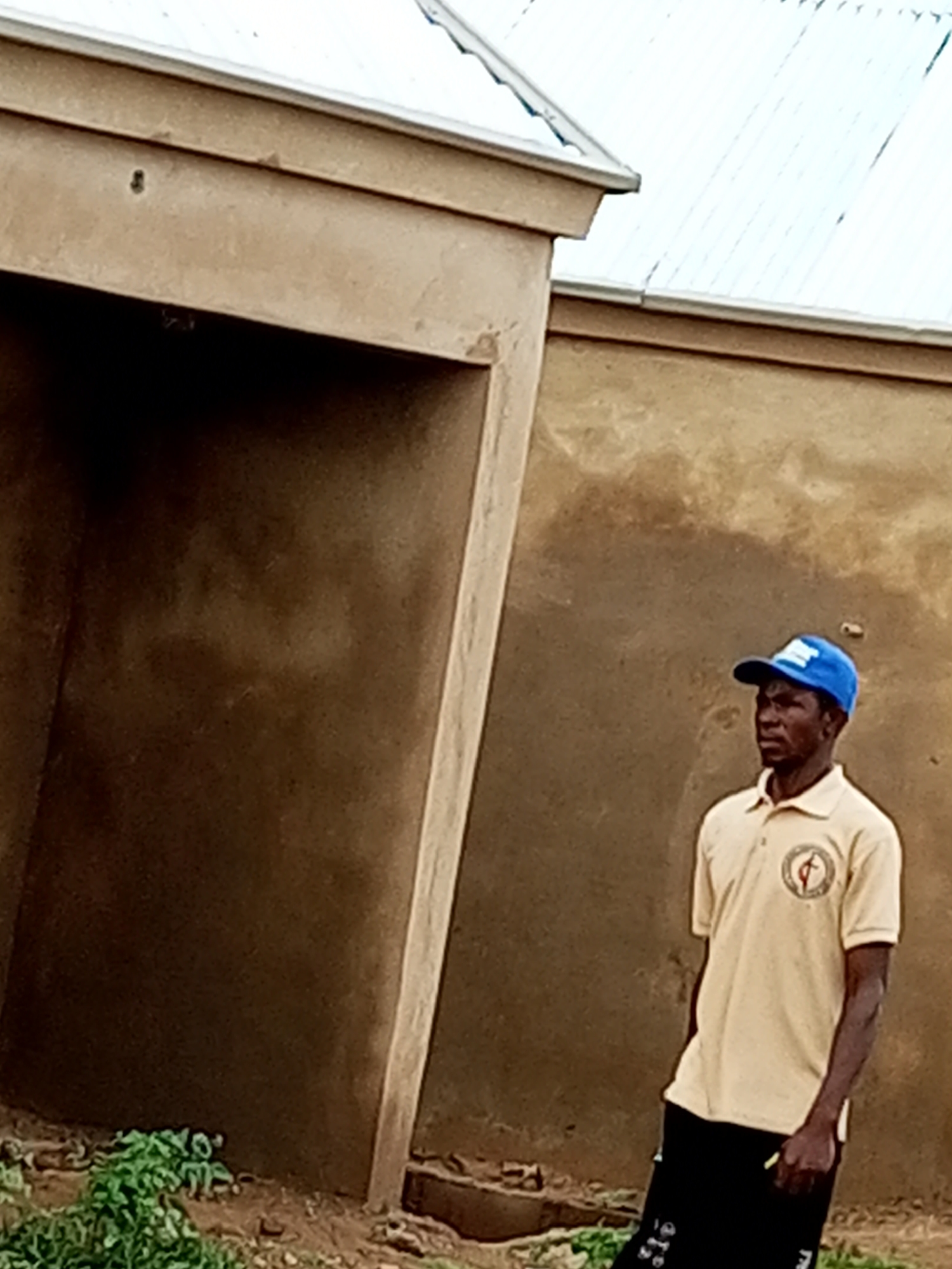 A medium shot shows a Black man standing outside. He is wearing a light tan polo shirt with a circular emblem on the left chest and a blue baseball cap. The man is looking to his right. Behind him is a textured, beige-colored wall and a section of a building with a white roof. In the foreground to the left, there is some green foliage and dirt. The man is standing in what appears to be a courtyard or an outdoor area next to a building.