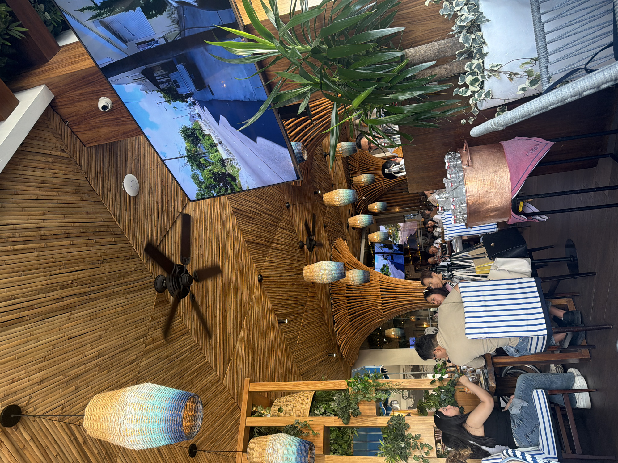 This image shows the interior of a restaurant, possibly in the city of Puebla, Mexico. The ceiling is made of bamboo and features a ceiling fan, hanging lamps, and a large screen displaying a video. There are people seated at tables with striped blue and white tablecloths and napkins, suggesting a dining establishment. The tables are set with glasses and what appear to be water pitchers. There is a copper container holding water in the foreground of the image. The restaurant has a decorative style with plants, and the overall lighting is warm. A security camera is visible on the ceiling.