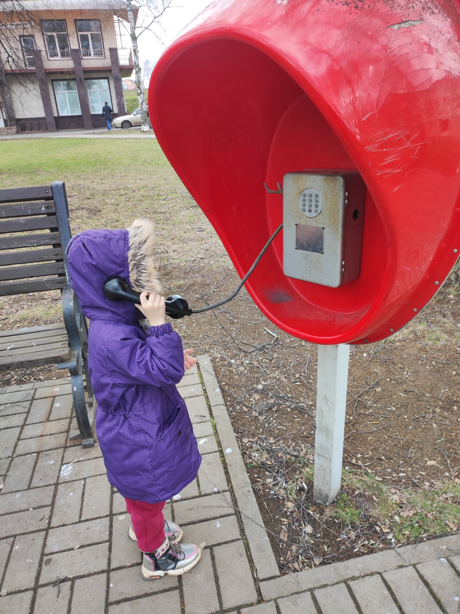 A young child wearing a purple winter coat with a fur-lined hood is standing on a paved area next to a red telephone booth. The child is holding the telephone receiver to their ear, as if engaged in a phone call. To the left of the child is a wooden bench. The background includes a building with windows, a patch of grass, a person walking, and a car. The scene appears to be set outdoors. The photograph was taken in the city of Votkinsk, Russia.