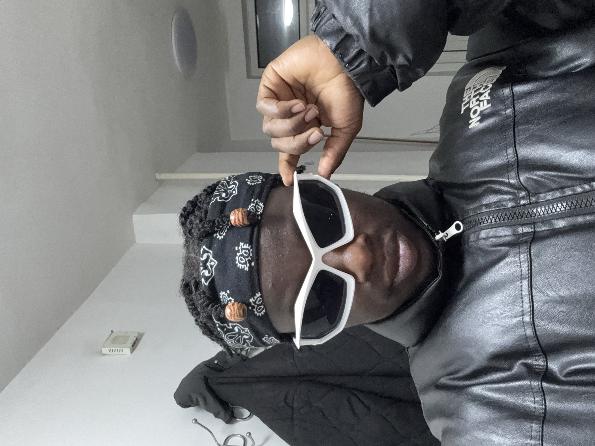 A close-up, tilted shot depicts a dark-skinned person wearing a black and white patterned bandana with brown beads, sunglasses with white frames, and a black leather jacket with "THE NORTH FACE" printed on the upper right side. The person's hand is visible, adjusting the sunglasses. The background shows a white wall with a circular ceiling light fixture and a small, rectangular device near the bottom left. A black garment hangs from a wall-mounted coat rack in the lower part of the frame.