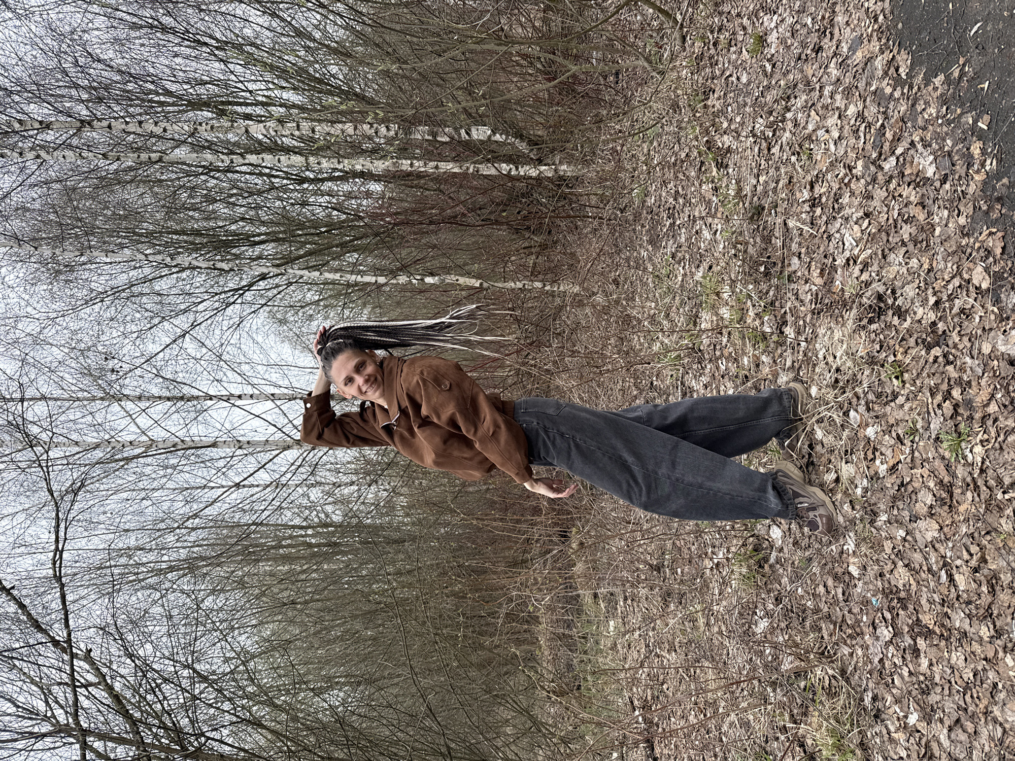 A woman poses outdoors in a wooded area. The woman is wearing a brown jacket and gray jeans. The environment includes trees with bare branches, a ground covered with dead leaves, and some evergreen shrubs. The location is specified as Rusanovo, Russia, but there are no specific details in the image to confirm this beyond the general appearance of the environment.