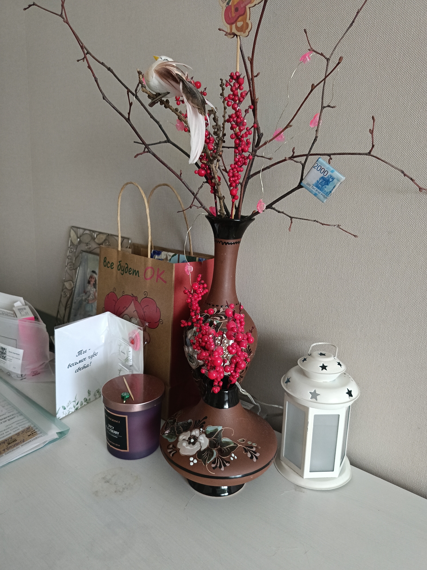 The image shows a still life composition on a white surface, possibly a table or shelf. The scene includes a brown vase adorned with red berries and artificial branches, featuring a decorative bird and a 2000 Russian Ruble bill attached to one of the branches. Alongside the vase, there's a white lantern with star-shaped cutouts, a purple candle, a framed photograph, a shopping bag with Russian text, and a greeting card also in Russian. The environment appears to be indoors, with a neutral-colored wall in the background. The text on the bag reads "Все будет ОК" (Everything will be OK). The card says "Ты - восьмое чудо света!" (You are the eighth wonder of the world!). The candle has the word "SPICY" and "BLACKBERRY" written on it. The bill is clearly labeled "ДВЕ ТЫСЯЧИ РУБЛЕ" (Two Thousand Rubles). There is no information to infer that the image was taken in Serpukhov, Russia.