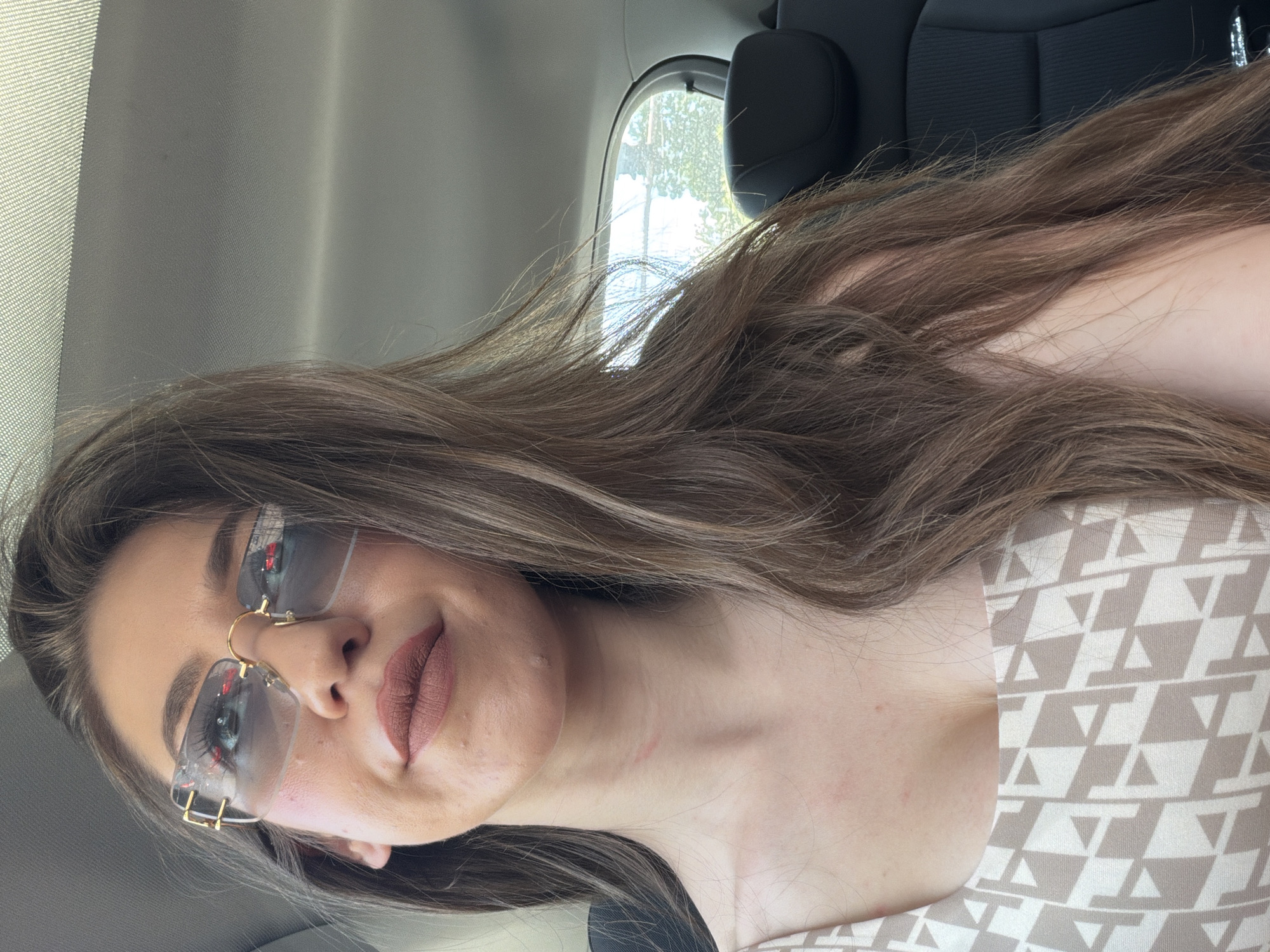 A young woman with long, brown hair and light-colored eyes is wearing oversized, rimless sunglasses with a gold frame. She has on a light brown and white geometric-patterned top. The woman is inside a vehicle, likely a car, with dark-colored seats visible in the background. The roof of the car and a side window showing trees and sky are also visible. Her makeup includes brown lipstick and mascara. She appears to be looking slightly upward and to her right.