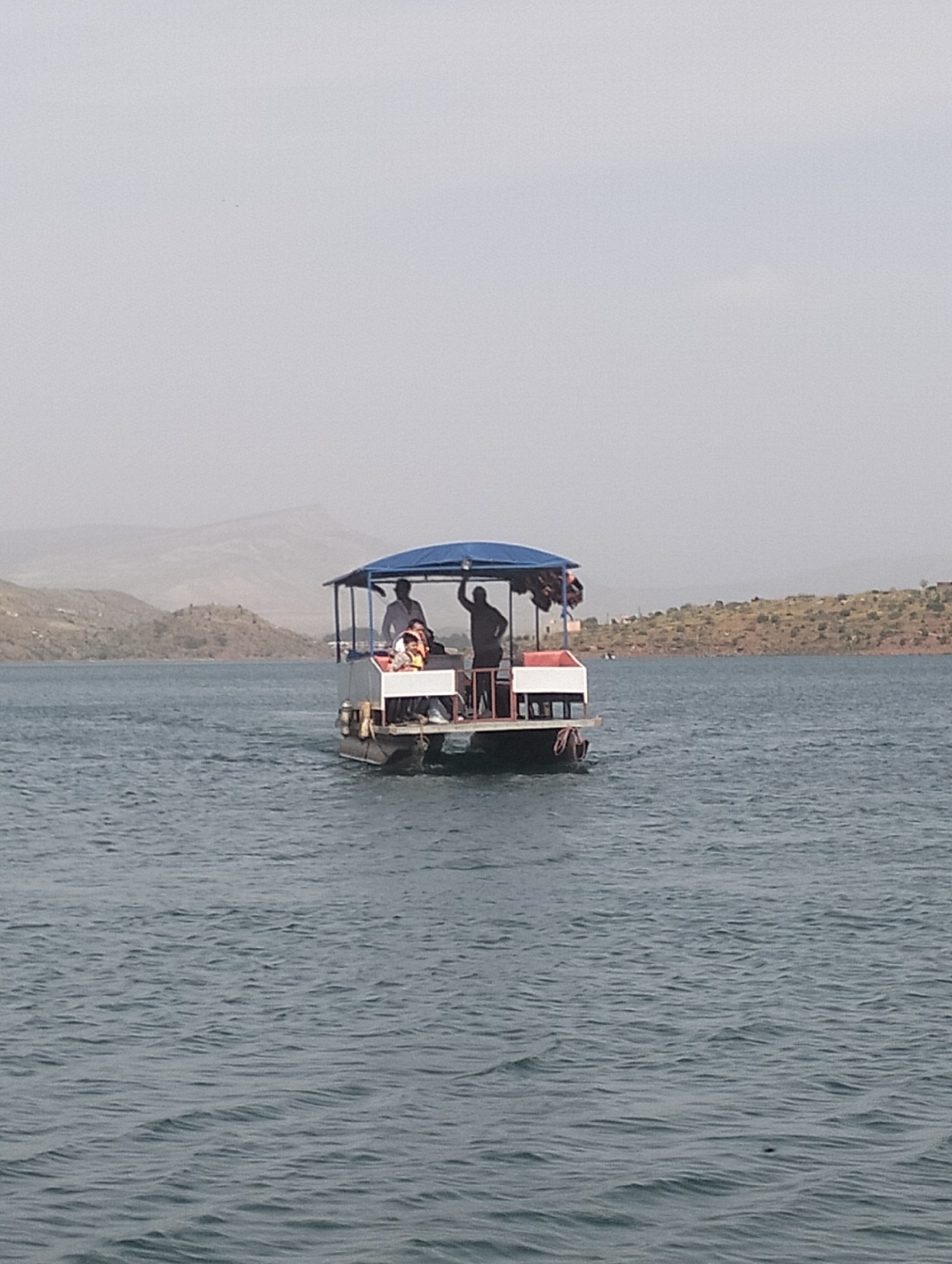 A medium shot captures a pontoon-style boat with a blue canopy navigating a large body of water, likely a lake or reservoir, in Bin El Ouidane, Morocco. Several people are visible on the boat; one person on the far right stands and waves their arm, while another person sits further back near the rear. At least two more individuals are partially visible towards the front of the boat, with one wearing an orange life jacket. The boat appears to be moving, leaving a slight wake behind it.
The sky is hazy and light grey, suggesting either an overcast day or atmospheric conditions reducing visibility. Distant, arid or sparsely vegetated hills and mountains frame the background, characteristic of the Moroccan landscape. The water is a deep blue-grey with gentle ripples across its surface. The overall impression is of a leisure activity, with people enjoying a boat ride in a scenic, rugged environment during daytime. No visible text is present in the image.