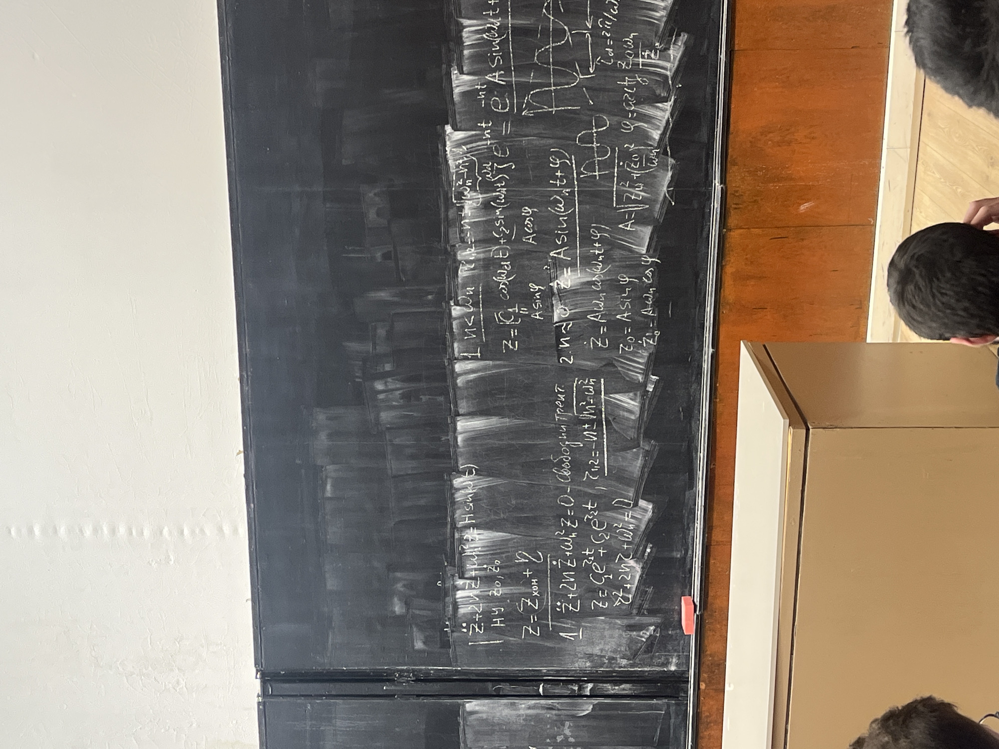 Here is a description of the image:
The image shows a section of a blackboard covered in mathematical equations and notes written in chalk. The blackboard is divided into sections, and the writing appears dense and complex, suggesting an advanced academic subject. Two people stand nearby looking at the blackboard. The environment appears to be a classroom or lecture hall in Sofia, Bulgaria based on other image metadata. The color scheme of the environment includes white and dark elements.