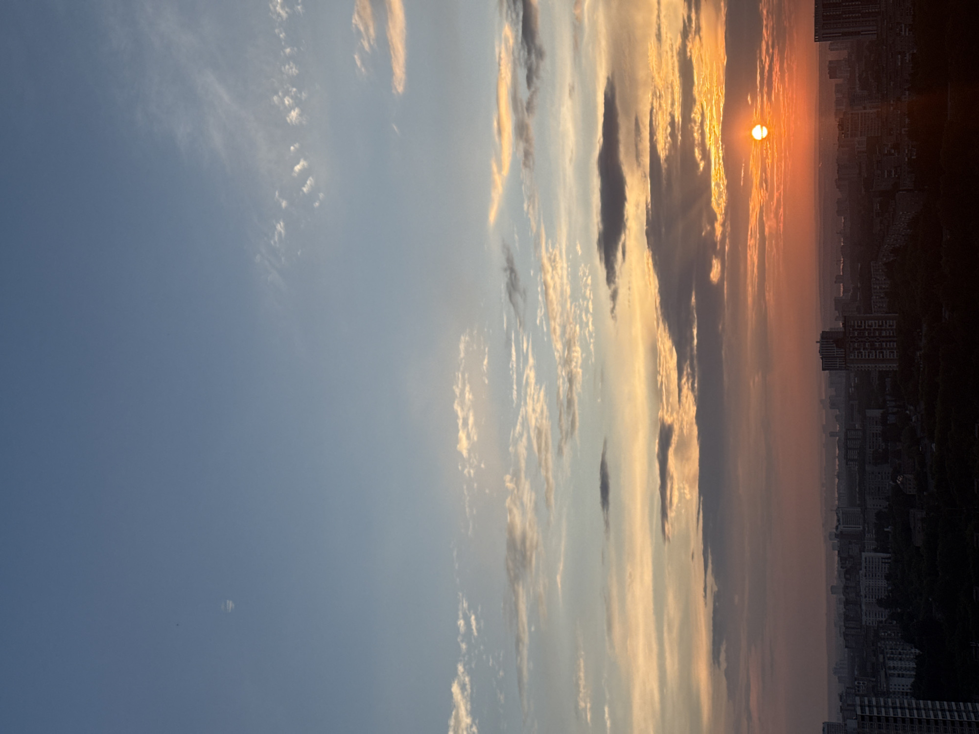 The image depicts a sunset over a city, likely Zarechnaya (Gamovskoe S/P), Russia. The sky transitions from a clear blue at the top to vibrant oranges and reds near the horizon. Wispy clouds are scattered across the sky, some catching the sunlight and appearing illuminated. The sun itself is a bright, orange orb, partially obscured by clouds, with visible rays of light emanating from it. Below the sky, a silhouette of a city skyline is visible. Numerous buildings of varying heights are present, with some taller, modern structures interspersed with lower, older buildings. The cityscape is largely dark, with the warm glow of the sunset casting a warm light on the upper portions of some buildings. The overall impression is one of a serene, urban landscape at dusk. No people or specific actions are discernible in the image.