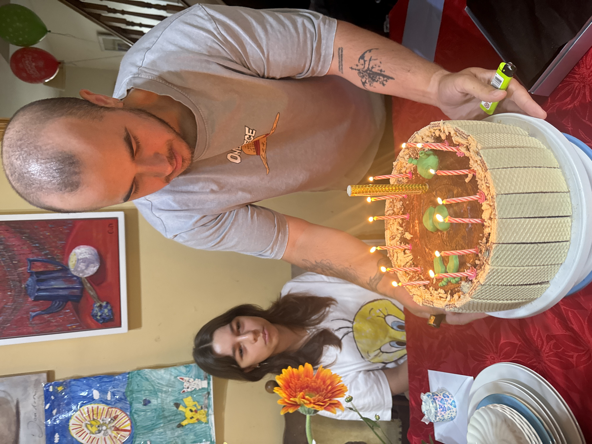 In a domestic setting, possibly a dining room in Waterford, Ireland, a man with a shaved head and a tattoo on his arm is lighting candles on a birthday cake. He is wearing a grey t-shirt with a "One Piece" logo and a distinctive orange hat graphic. Beside him, a young woman with long brown hair, wearing a white t-shirt with a Tweety Bird graphic, watches the cake. The cake is decorated with chocolate frosting, green icing shapes, and numerous lit candles. The table is covered with a red, patterned tablecloth. On the table, there are stacks of plates, a small decorative item, and a lighter in the man's hand. In the background, a wall is adorned with several framed art pieces, and two balloons, one red and one green, are visible near a staircase.