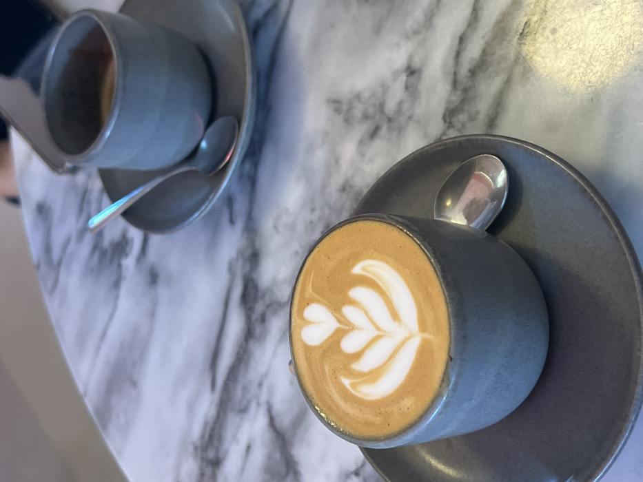 The image displays a close-up, high-angle view of two coffee servings positioned on a white marble-patterned table featuring dark gray veining. In the foreground, a gray ceramic cup, approximately half-filled, contains a light brown coffee beverage adorned with white foam latte art forming a stylized tulip or multiple heart design. This cup rests on a matching gray ceramic saucer, with a small silver spoon situated to its right. In the blurred background, a second, similar gray ceramic cup holds a darker brown liquid, likely an espresso, also placed on a matching gray saucer with a small silver spoon. Partially visible on the table in the background, near the second cup, is a smartphone and a segment of a person's hand. The specific location within Lordelo do Ouro e Massarelos, Portugal, is not discernible from the visual information provided.