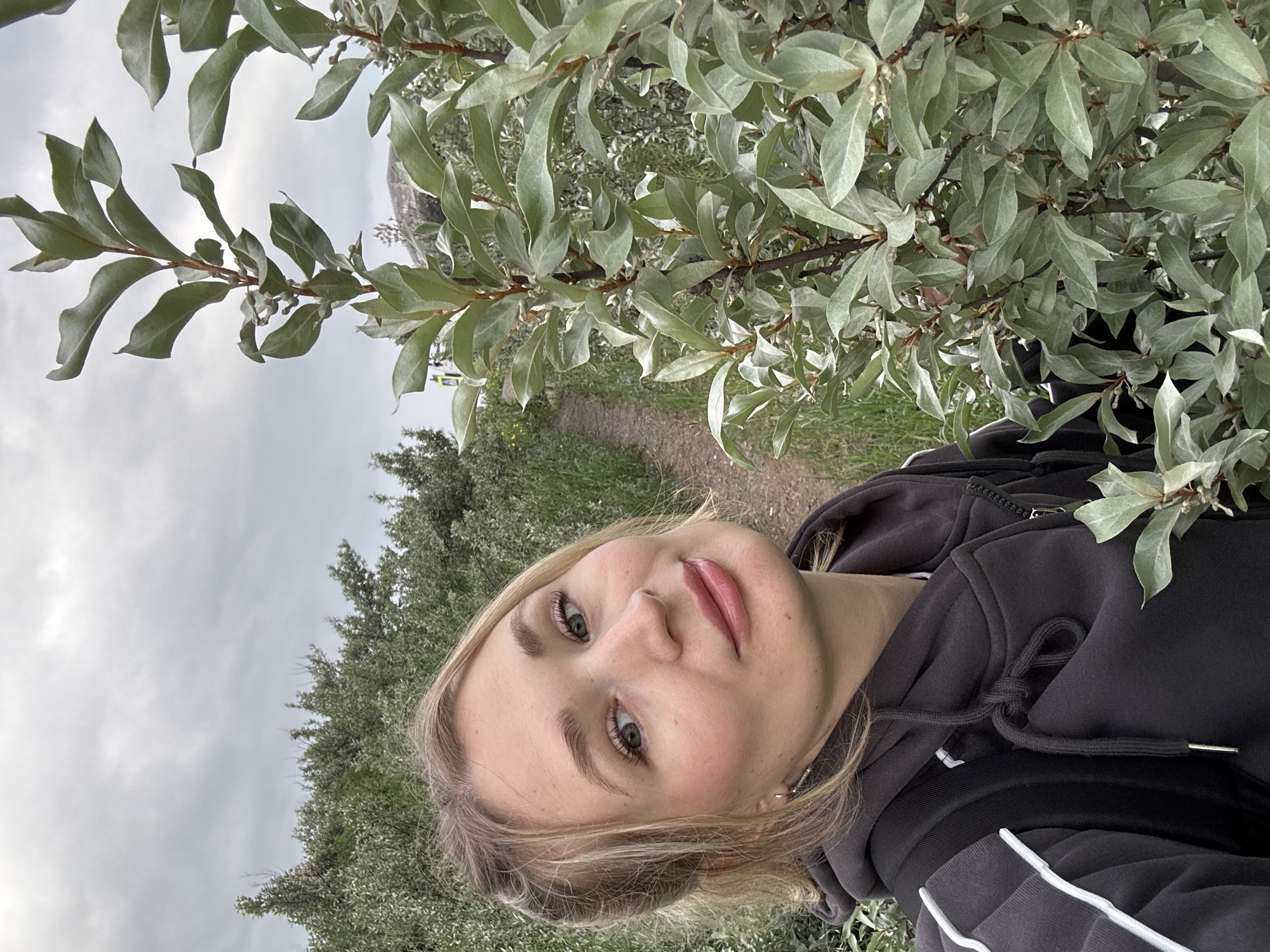 The image shows a young woman with blonde hair tied back, taking a selfie outdoors. She is wearing a dark grey hooded sweatshirt and a backpack. She has light-colored eyes, fair skin with blush on her cheeks, and pink lipstick. The background is a natural setting with a path winding through greenery. There are trees and bushes on either side of the path, and the sky is overcast. The woman is positioned in the foreground, with a plant prominently in front of her on the right side of the frame. The location is not identifiable as Chaikovsky, Russia, or any specific city.