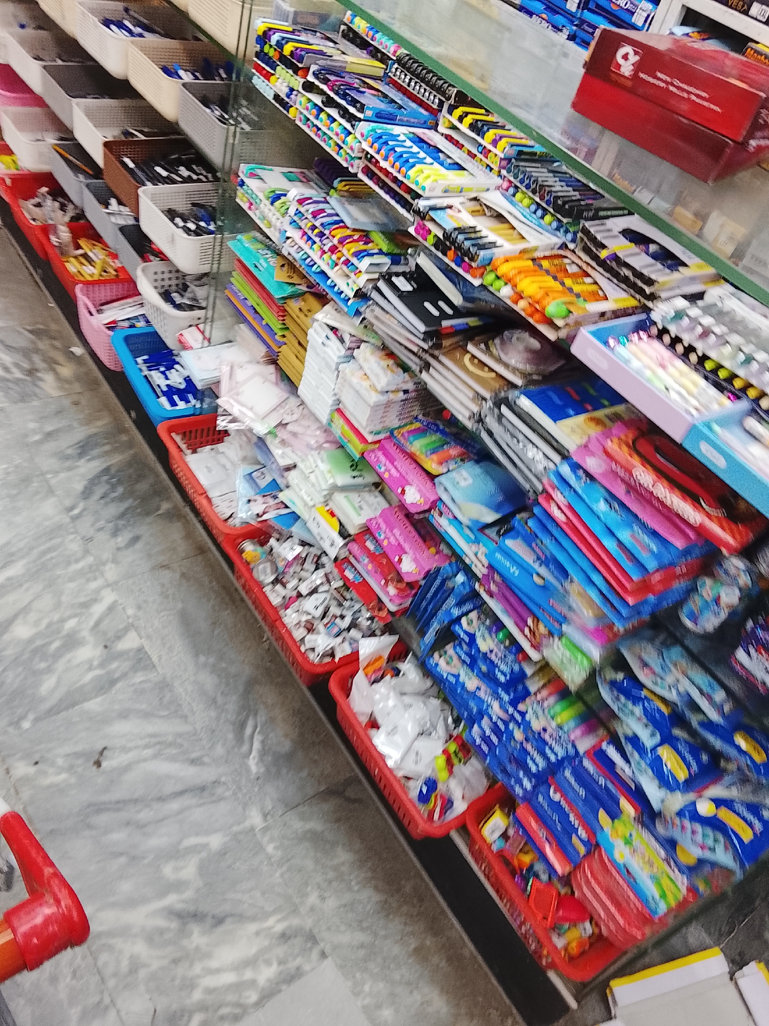 The image shows a retail display in Gulgasht Colony, Pakistan. The display consists of a multi-tiered shelf showcasing stationery supplies. The items include pens, notebooks, and various art supplies, organized in rows and plastic baskets. The floor is light-colored tile. A portion of a red shopping cart handle is visible in the foreground.