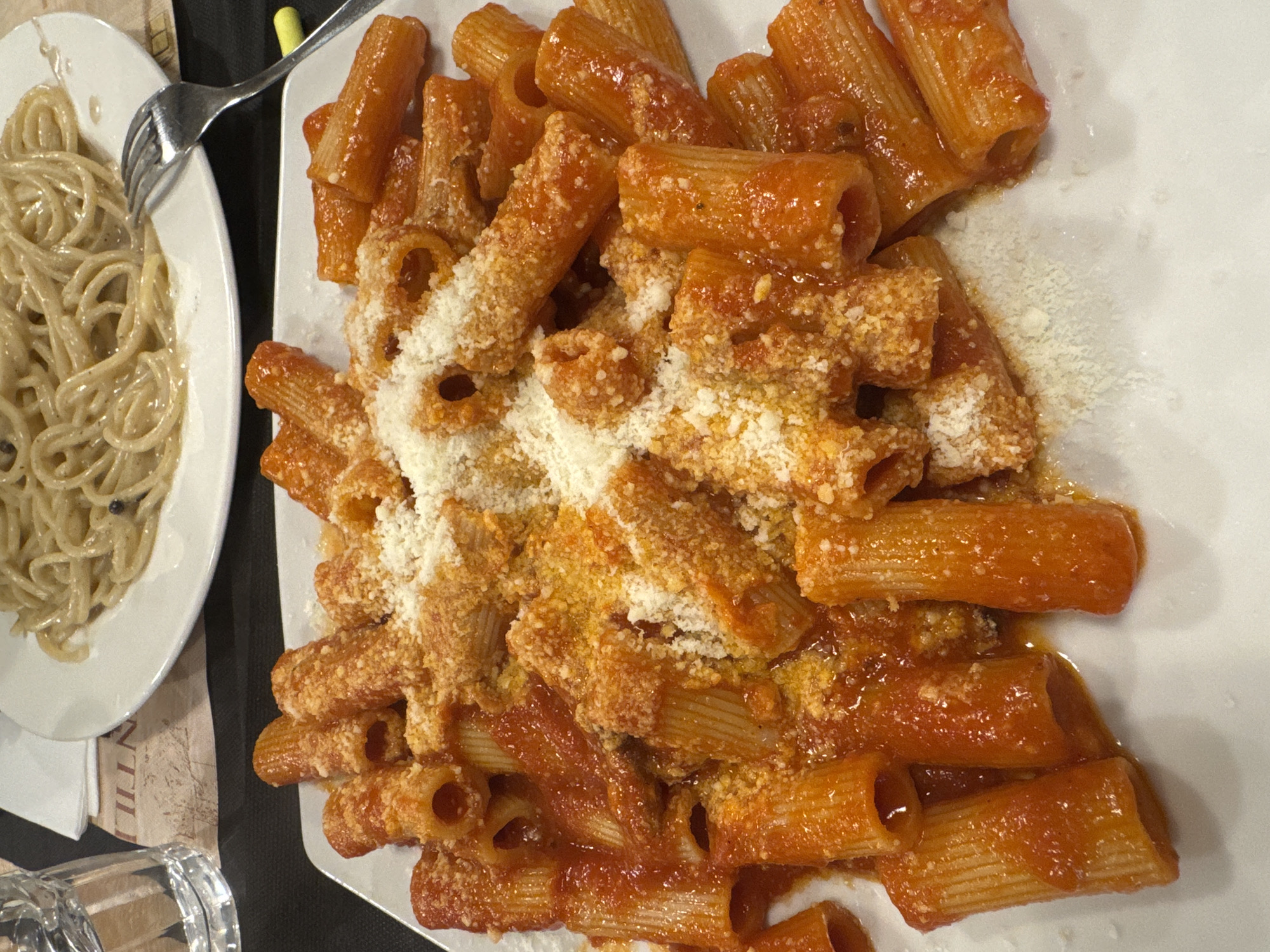 This image captures a close-up, overhead view of a dining table displaying two pasta dishes in what appears to be a restaurant in Rome, Italy. In the foreground, a rectangular white plate is filled with rigatoni pasta, coated in a rich, reddish-orange tomato-based sauce. A substantial amount of finely grated white cheese, likely Pecorino Romano or Parmesan, is generously dusted over the pasta, along with some visible yellow-orange seasoning. In the upper background, out of focus, a portion of a round white plate holds spaghetti prepared with a light-colored, creamy sauce, featuring distinct black pepper flakes, characteristic of Cacio e Pepe. A silver fork rests on the right edge of this plate, and a clear glass tumbler containing water is partially visible on the upper left. The table surface features a dark placemat or tablecloth underneath, and a patterned beige paper placemat, with the partial text "NTILLI" visible, suggesting a restaurant or brand name. No individuals are present in the frame.