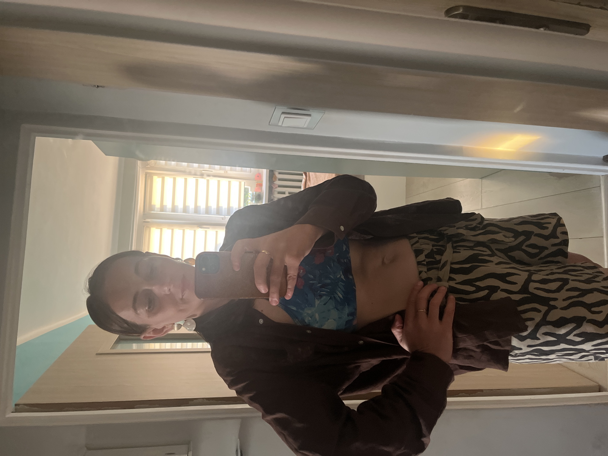 A woman is standing in a doorway and taking a mirror selfie. She is wearing a brown open shirt over a blue floral bra top and tan and black zebra-print shorts. She has on large, circular earrings and is wearing a ring on her left hand. She is holding a brown phone in her right hand, which is angled to capture the reflection. Her left hand is on her hip. The environment appears to be a home interior, with a doorway leading to a room with a window and blinds. The floor is tiled.