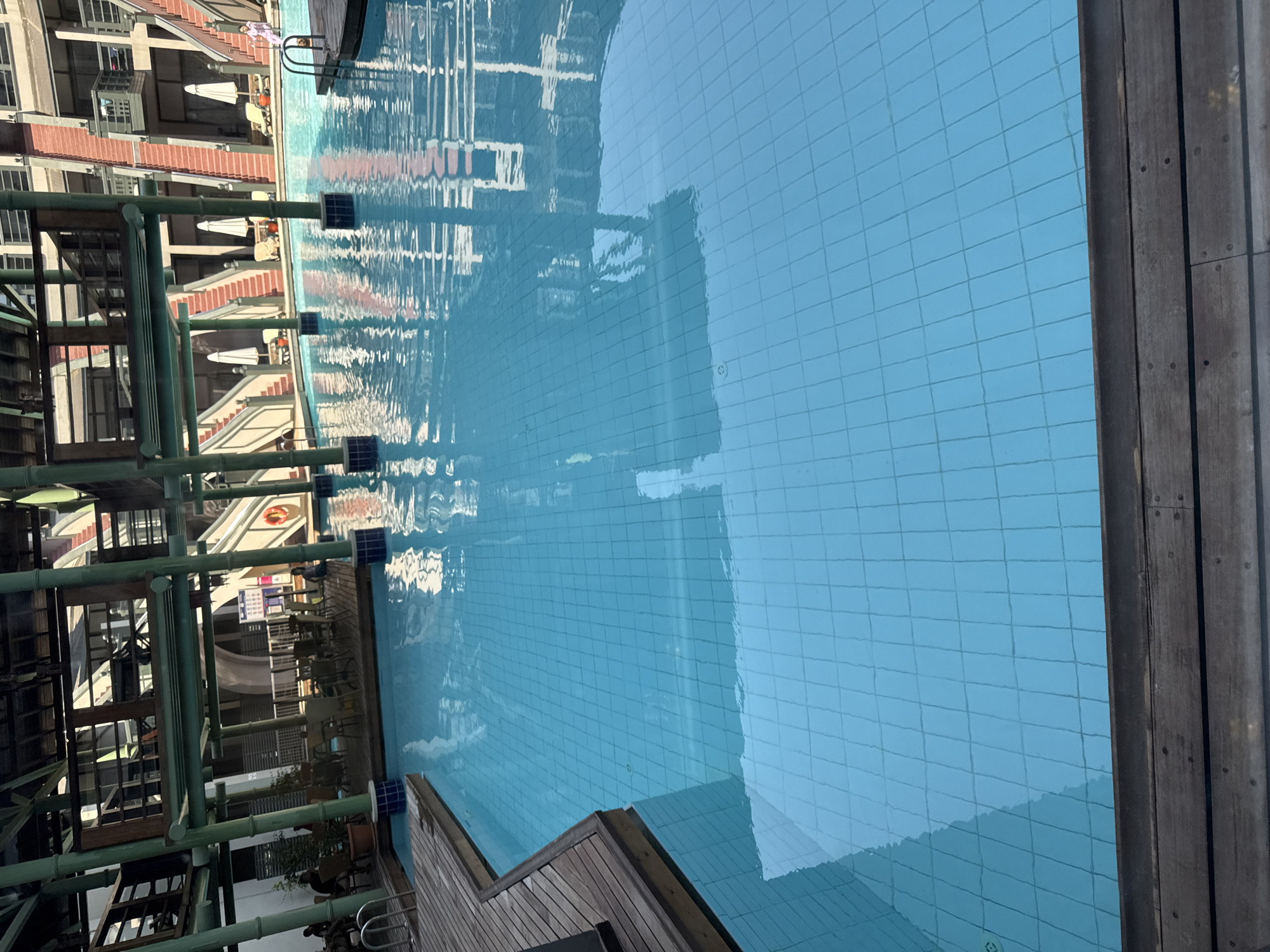 The image depicts a swimming pool area, likely within a resort or hotel complex, in the city of Aksu, Turkey. The pool is large and filled with clear blue water, tiled on the bottom, with reflections of the surrounding architecture. In the background, there are multiple levels of buildings with open walkways, staircases, and balconies. Several white umbrellas and lounge chairs are visible near the pool. There are a few people in the distance, some seated and others standing. A red life preserver is attached to a pole on the right side of the pool. The foreground shows a wooden deck bordering the pool. The overall scene suggests a leisure and recreational environment.