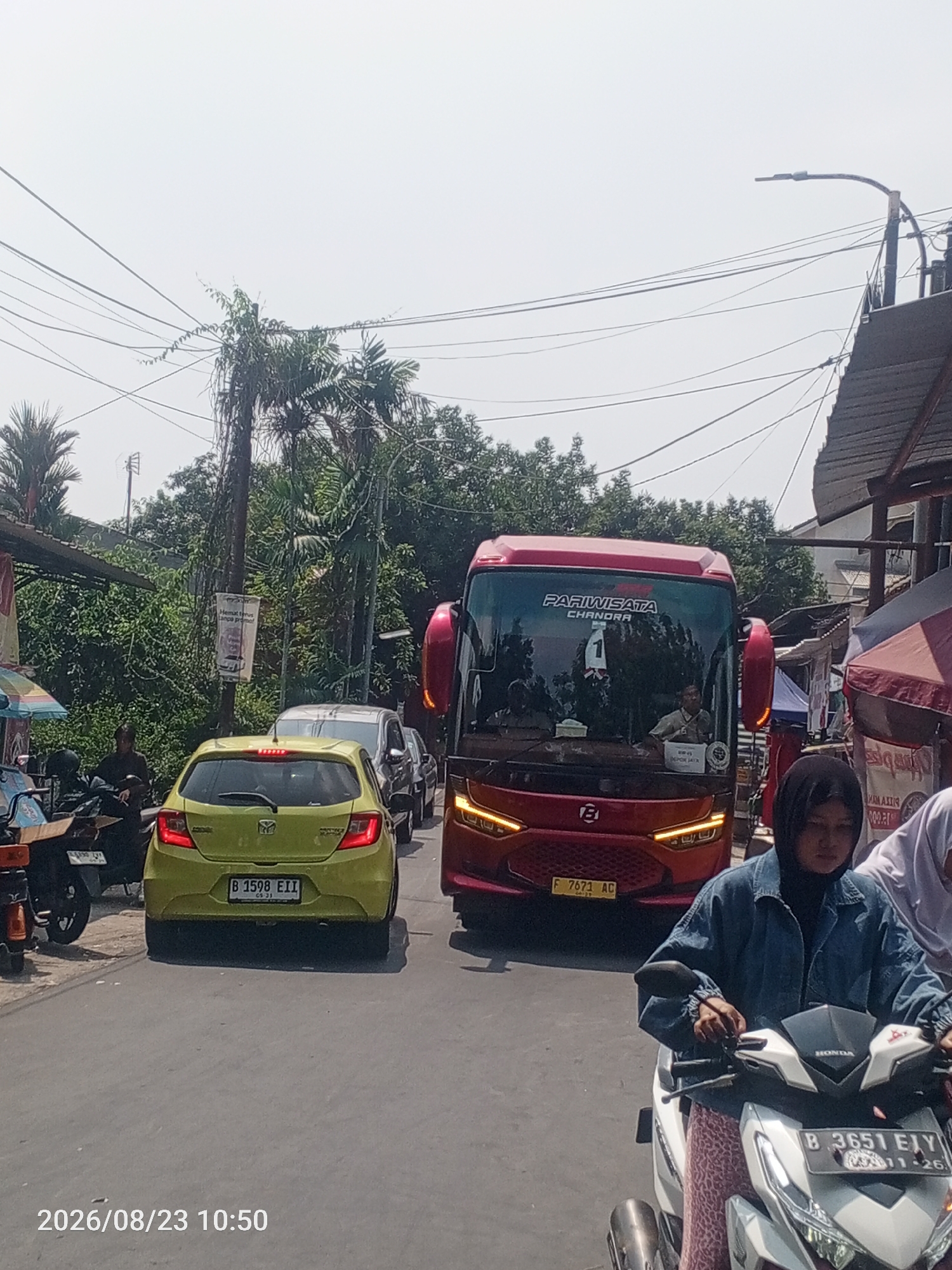In Depok, Indonesia, a daytime traffic scene unfolds on a paved road lined with lush green trees and electrical wires overhead. In the foreground, a person on a white Honda scooter, wearing a blue denim jacket and a dark hijab, is stopped at an intersection. The scooter's license plate reads "B 3651 EIY". Behind the scooter, a yellow car with license plate "B 1598 EII" is also stopped. Further down the road, a large red bus with "PARIWISATA CHANDRA" printed on its windshield is visible. The bus has its headlights illuminated. Other vehicles, including a silver car, are also present in the traffic jam. The environment suggests a typical urban setting with a mix of vehicles and foliage. The timestamp at the bottom left corner indicates the image was captured on August 23, 2026, at 10:50 AM.