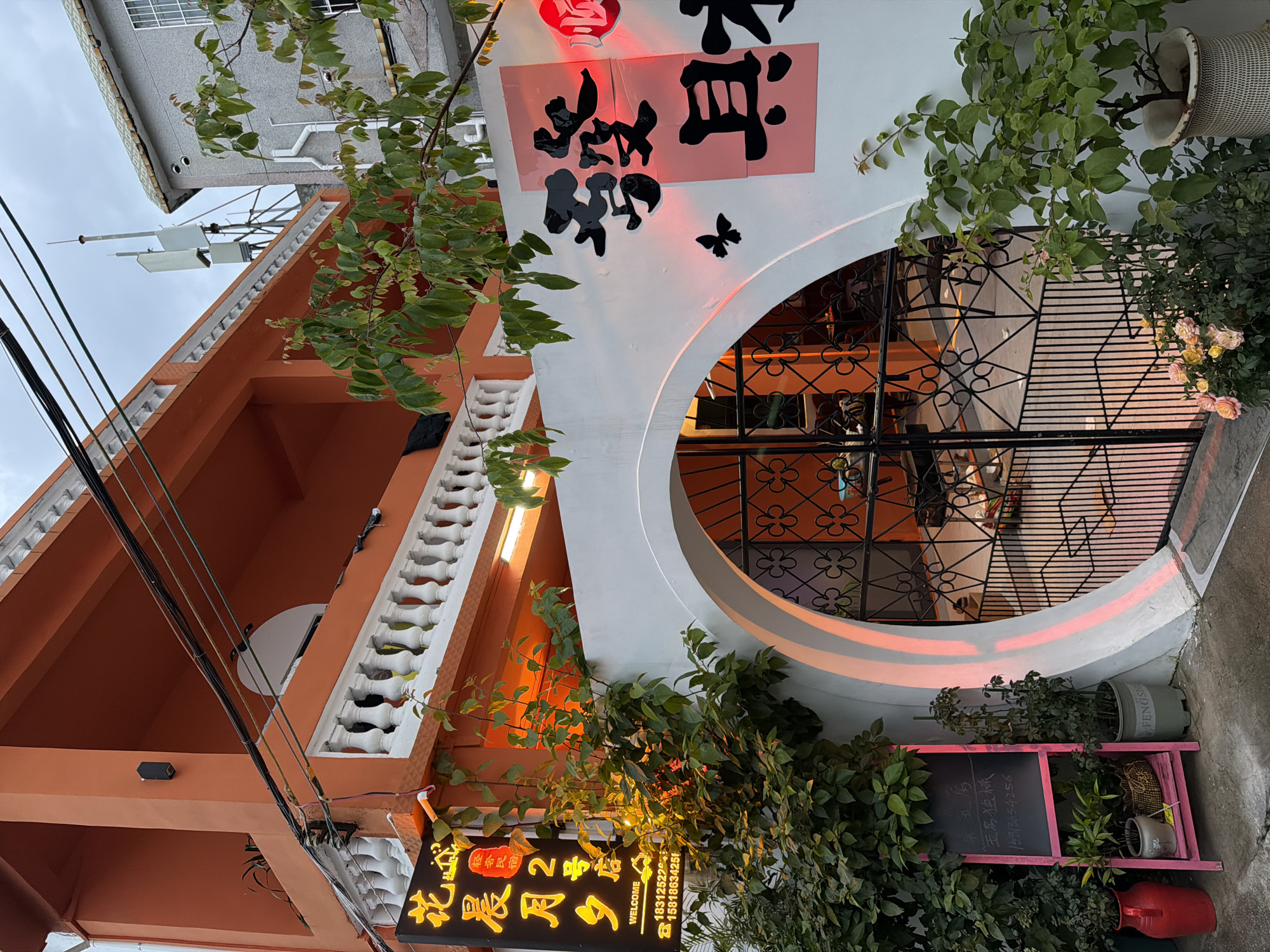 The image depicts the exterior of a business establishment, likely in the city of Longgang District, China, characterized by its vibrant orange facade. A prominent feature is a large, arched entryway with ornate black metal gates, leading into what appears to be a commercial space. To the left of the entrance, a black sign with yellow Chinese characters and the word "WELCOME" in English is illuminated, along with two phone numbers. Adjacent to the sign is a pink A-frame chalkboard displaying Chinese text, and below it are several potted plants. The white wall surrounding the archway is decorated with large black Chinese characters and a small black butterfly silhouette. Green foliage from nearby plants spills over the wall and frames the entrance. The building features a white balustrade on the upper level. Overhead, electrical wires crisscross the sky, and a cell tower is visible in the background. The lighting suggests it is either dusk or an overcast day, with warm light illuminating parts of the signage and the interior visible through the gate.