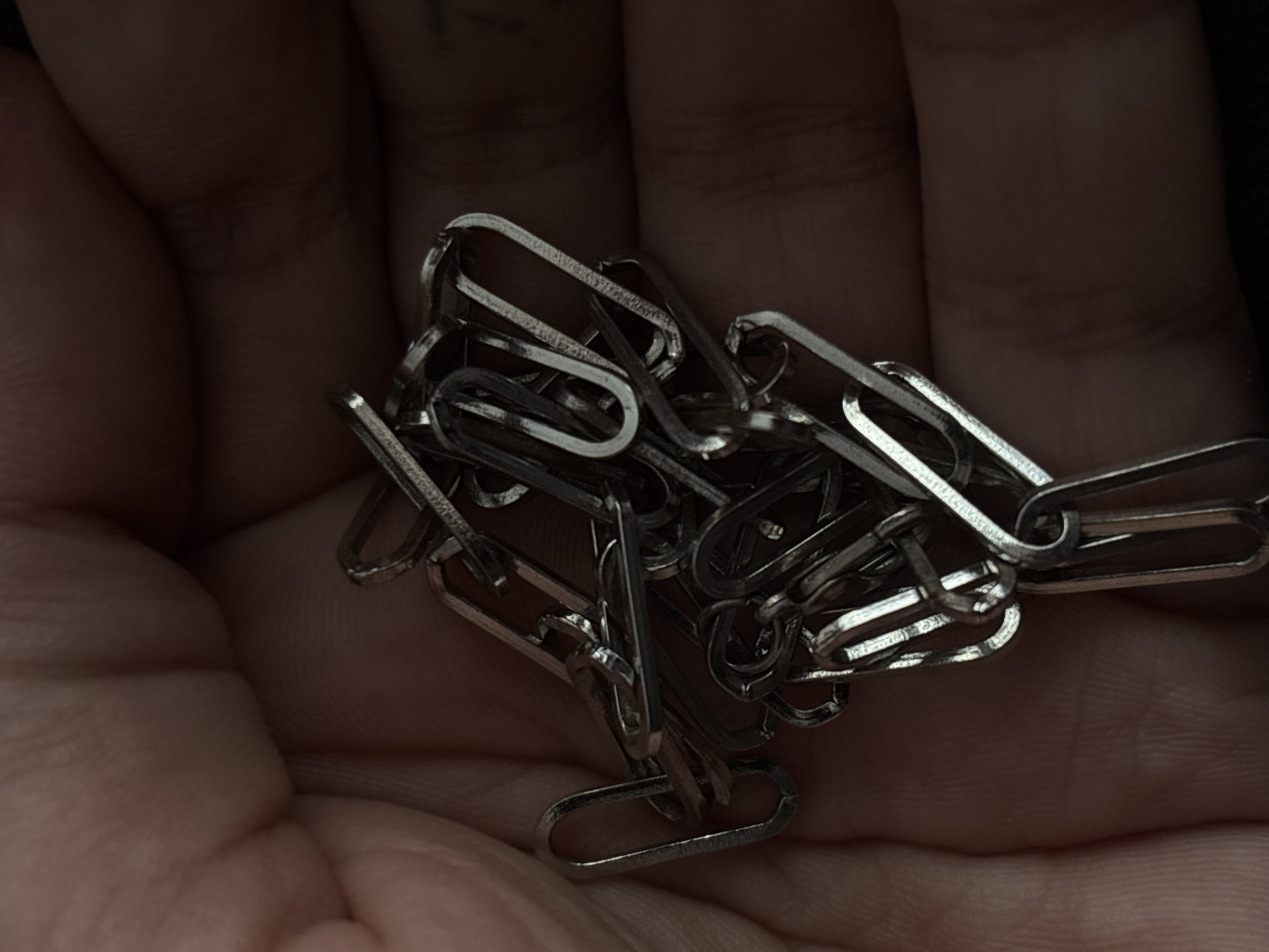 A close-up, overhead shot captures a jumble of silver paperclips intertwined and held in a person's cupped hand. The hand, which appears to be that of an adult, is flesh-toned and shows visible skin texture and faint creases, suggesting it is the thumb and forefinger region. The lighting is dim, creating shadows that obscure details of the background and the finer points of the paperclips. The scene is devoid of any discernible environmental context, making it impossible to ascertain its location within Saint Petersburg, Russia, or any other city.
