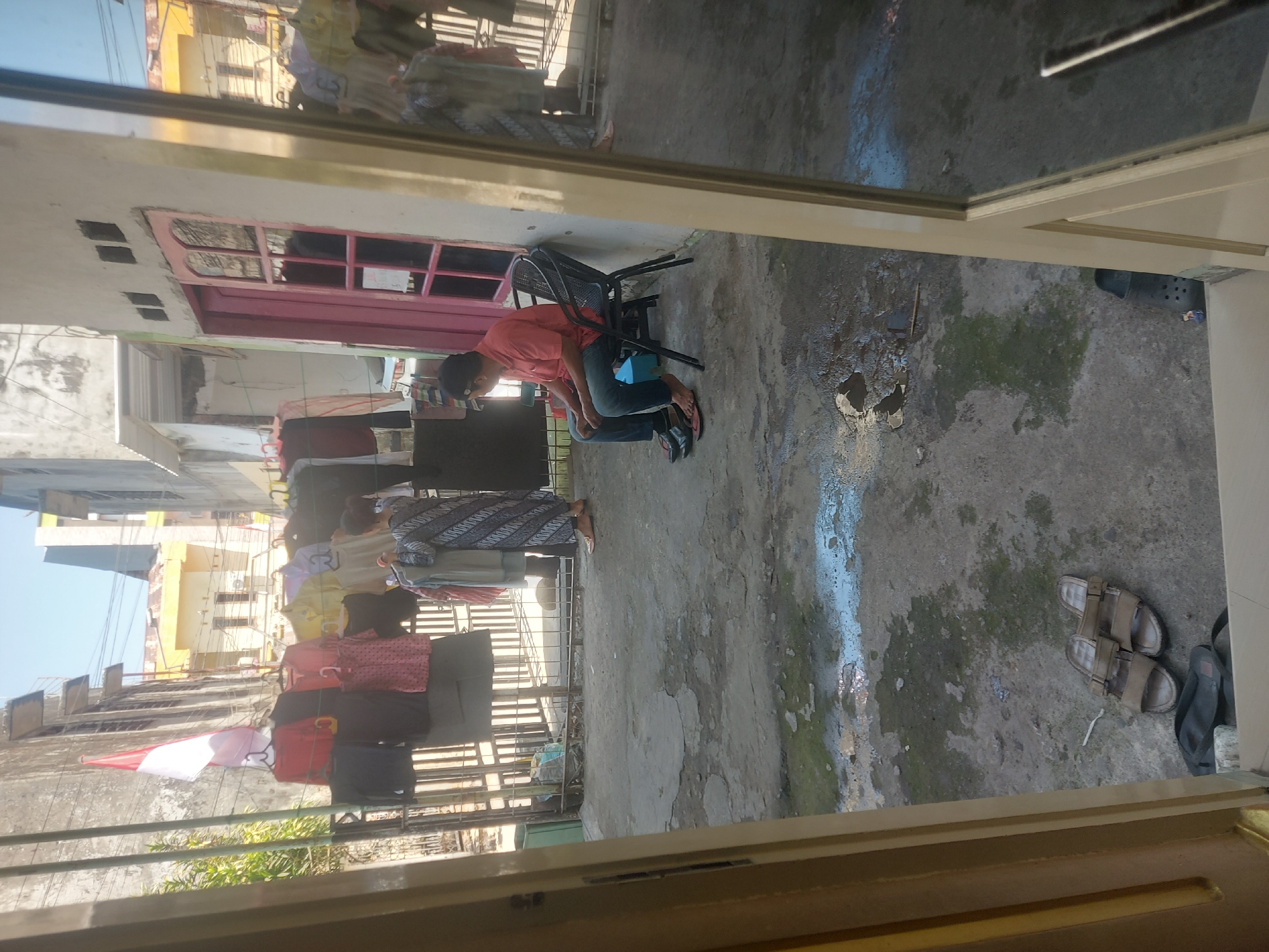A medium shot captures a residential exterior in Makassar, Indonesia, under a clear, bright sky. The scene is viewed through an open doorway, with a wall and door frame on the left and a reflective glass door on the right. In the foreground, a pair of brown sandals rests on the concrete ground near a dark bag. The ground itself is textured concrete, showing patches of dampness and some debris, indicating potential recent rain or water use.
Further into the scene, a man in a coral-colored short-sleeved shirt and blue jeans sits on a black outdoor chair, facing away from the camera and towards the right. He wears flip-flops. Behind him, a fence runs horizontally across the middle ground. A clothesline strung across the fence displays a variety of hanging garments, including red, black, yellow, and white shirts, and a pink patterned dress. An Indonesian flag, red and white, flutters from a pole near the fence.
To the right of the man, a pink door with glass panes is visible. Beyond the fence, the background shows multi-story buildings with yellow and white facades, characteristic of urban Indonesian architecture. The reflection in the glass door on the right provides a partial view of the same scene, including more hanging clothes and a person in a patterned dress standing near the fence. The overall impression is a typical residential setting in a bustling Indonesian city, with laundry being air-dried outdoors.