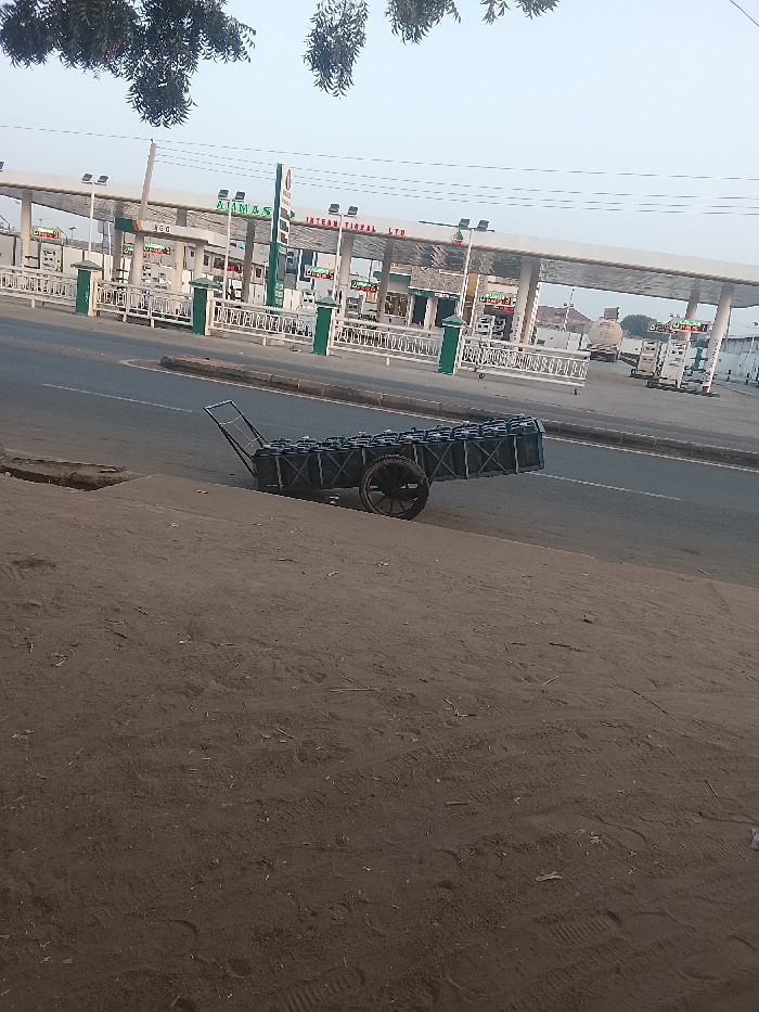 A two-wheeled cart, packed with black containers, rests on a curb alongside a road. In the background, a sprawling gas station complex with numerous pumps and signs is visible. The sky is overcast, and bare trees are in the top left corner of the frame. The foreground is dominated by a dirt or sandy surface, possibly a roadside verge.