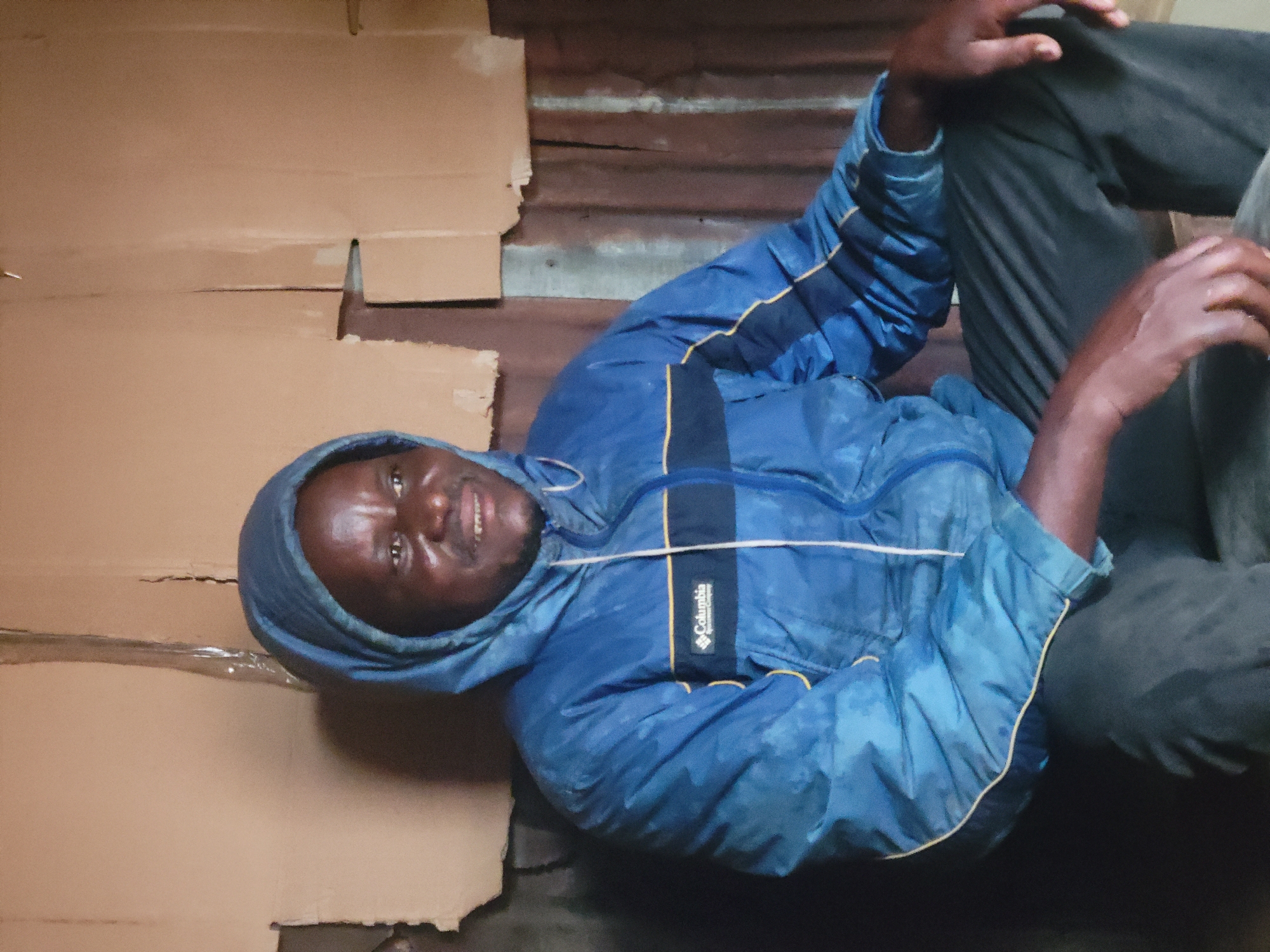 A medium shot captures a Black man in his late 30s or early 40s, sitting down and looking to his right, with a slight smile. He is wearing a blue hooded jacket with yellow trim and the "Columbia Sportswear Company" logo visible on the left chest. His skin appears to be dark, and he has short, dark hair. He is also wearing dark pants.
The background consists of rough, uneven walls made of what appears to be corrugated metal and cardboard. The lighting is dim, casting shadows on the walls and the man's face. The overall impression is of an indoor space that is sparsely furnished and somewhat dilapidated. The scene is likely a residential interior, possibly in Madalla, Nigeria, given the context.